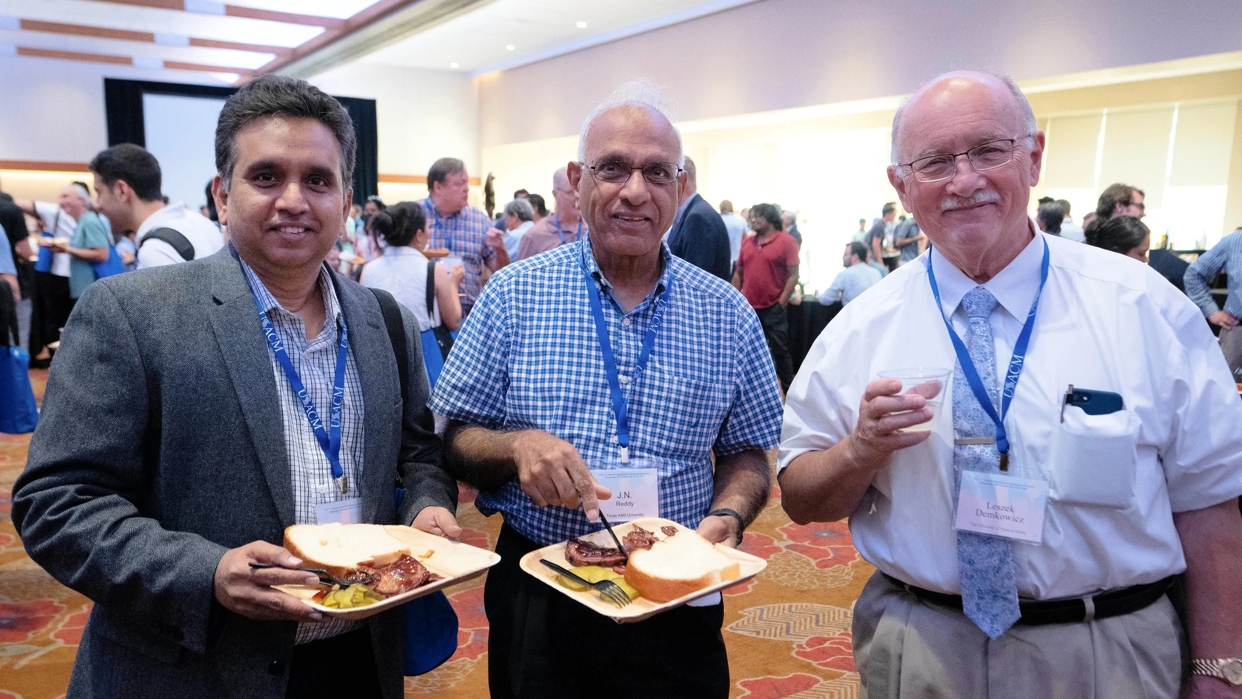 Three men in business attire and name badges, holding trays with food and drinks, at a conference or networking event with many people in the background.
