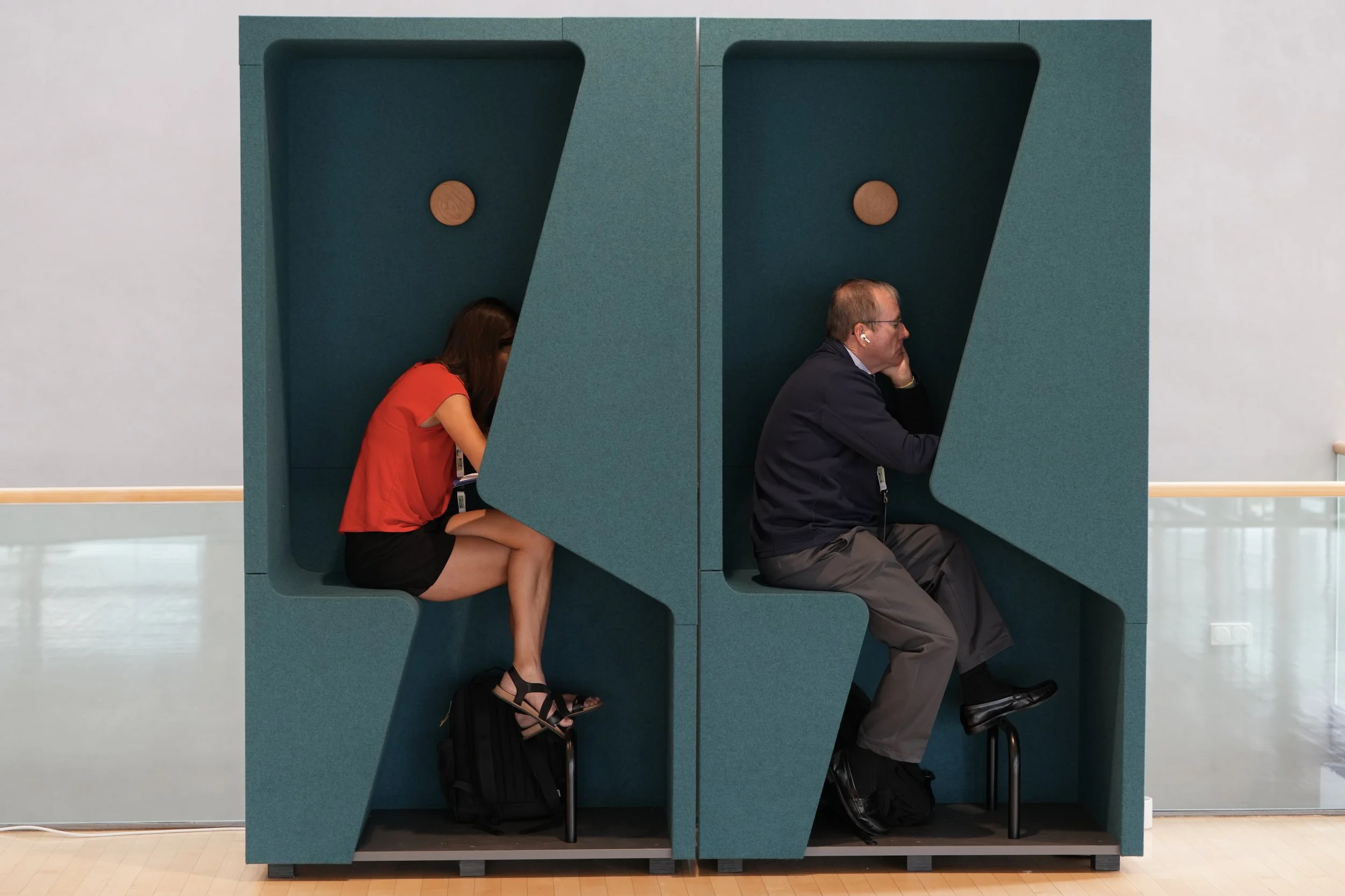 Two people sitting in a soundproof booth at an airport, each focused on a device, with their luggage underneath. The booth is teal with two separate compartments, each with a wooden accent on the interior wall.
