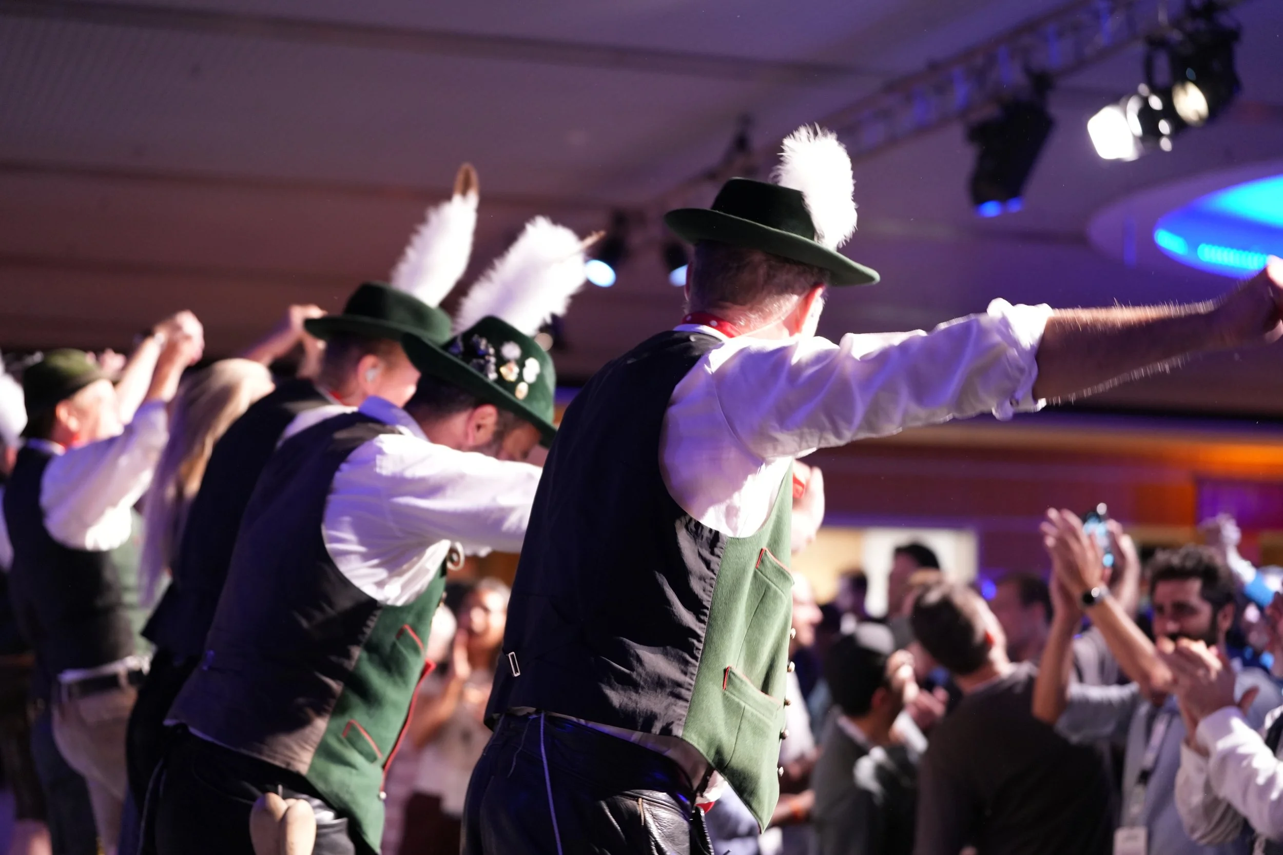 People dressed in traditional costumes with green vests, white shirts, and hats with feathers, performing a dance or ceremony at an indoor event with stage lighting.