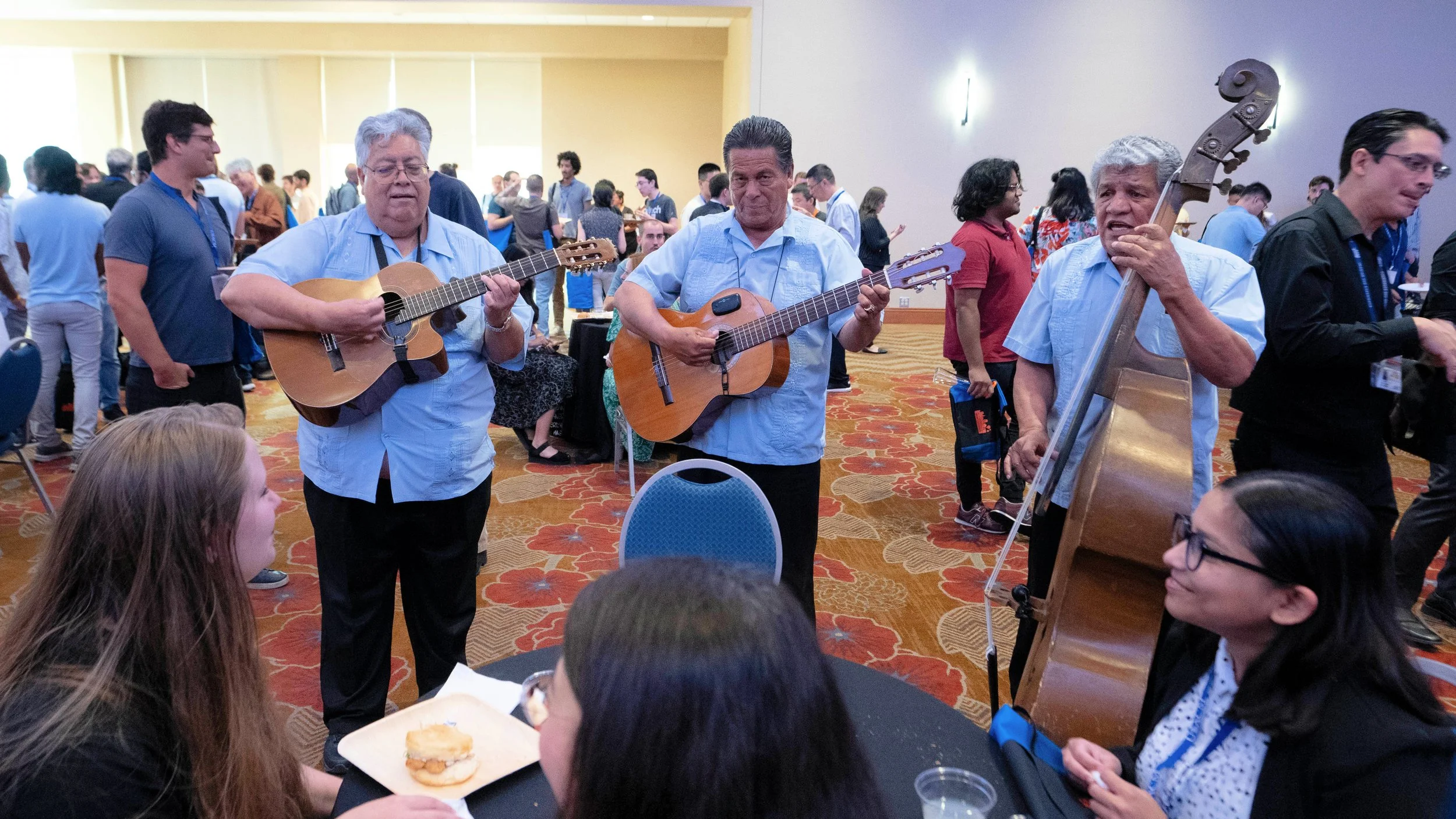 Group of four men playing acoustic guitars and singing at a social event, with a crowd of people standing and sitting in the background.