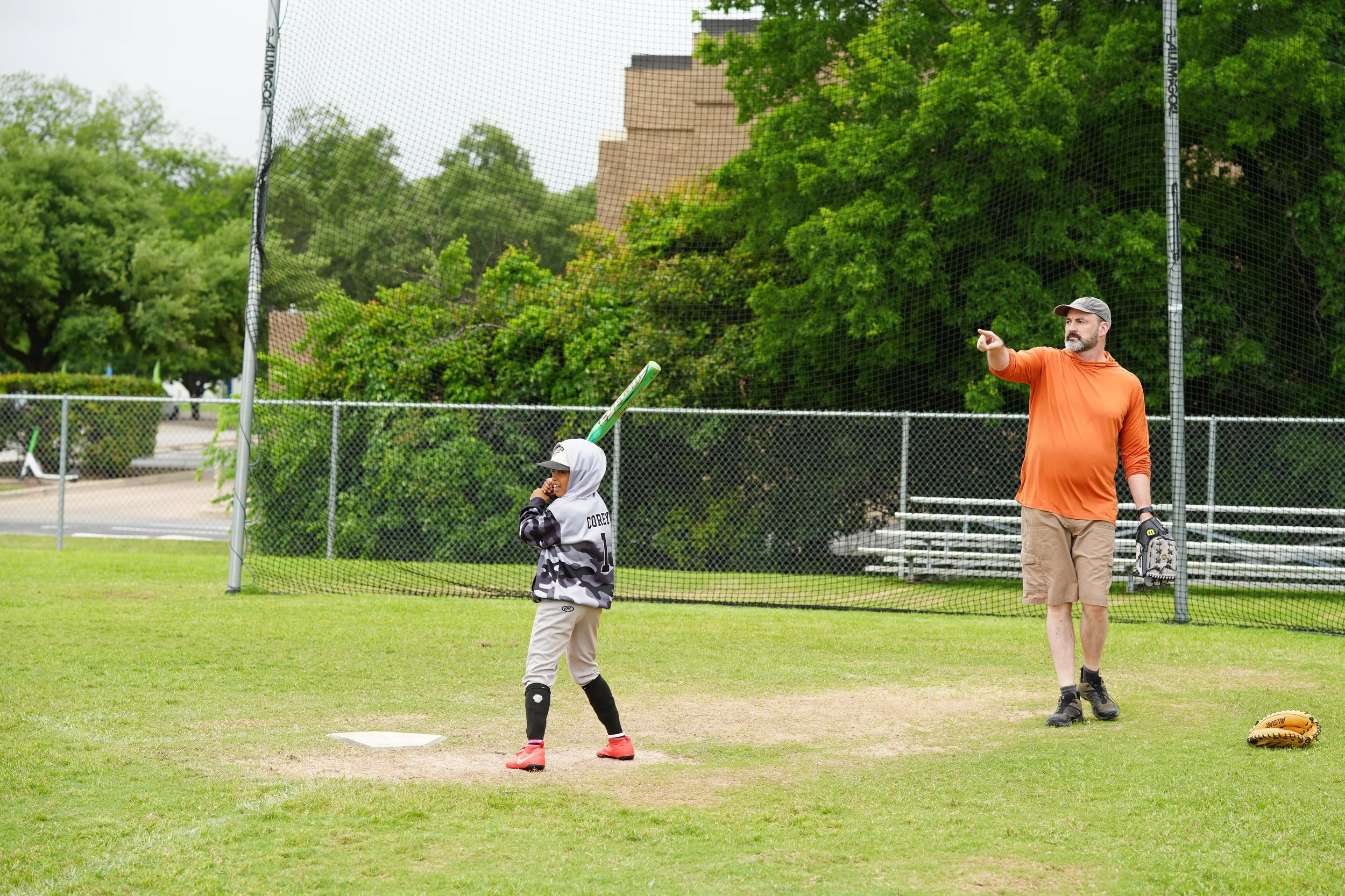 A man and a young girl on a baseball field, with the man instructing or giving directions and the girl holding a baseball bat, behind a protective netting, with trees and a building in the background.