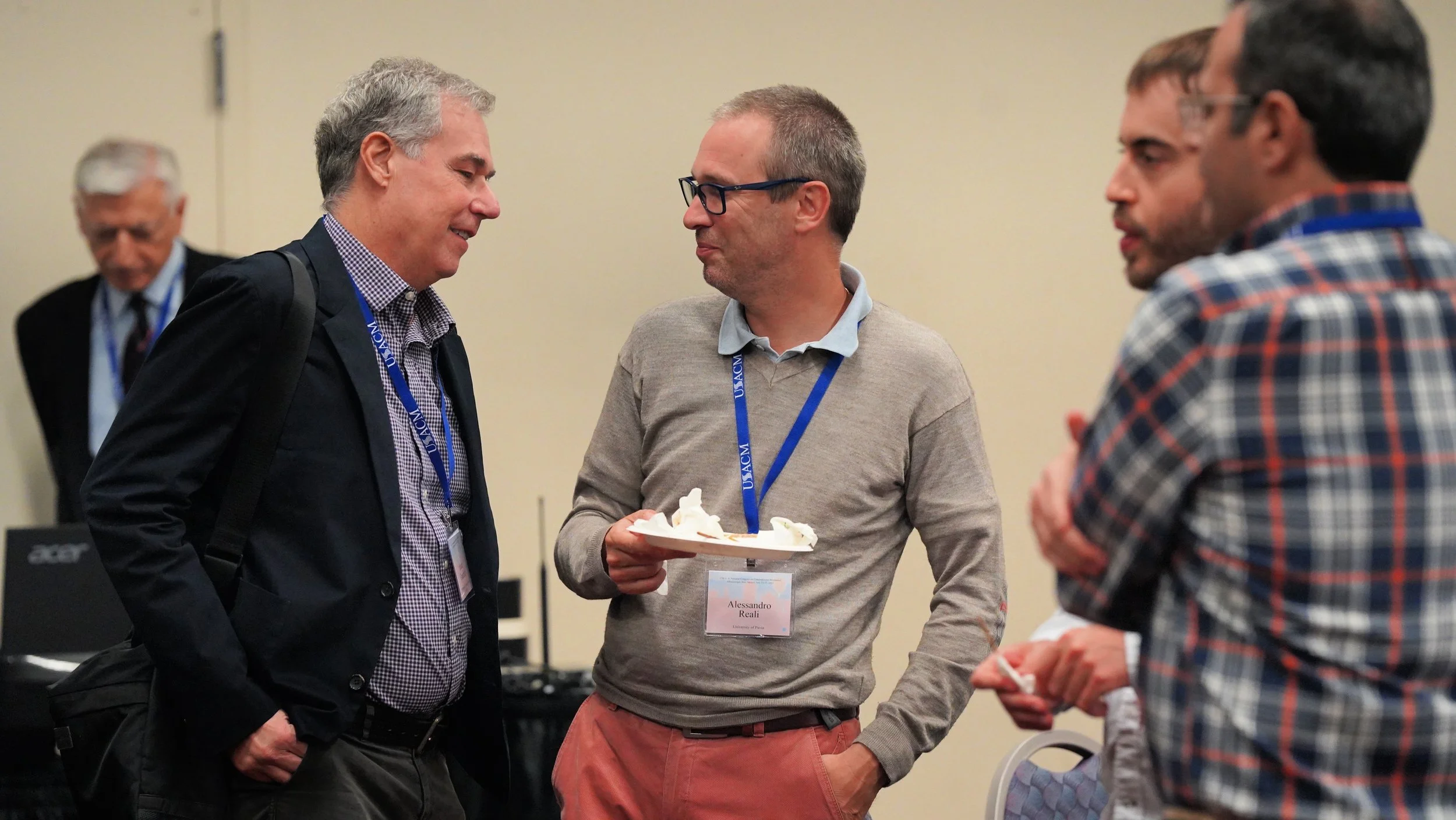 Four men engaged in conversation at a conference, one holding a plate with dessert, all wearing conference badges.