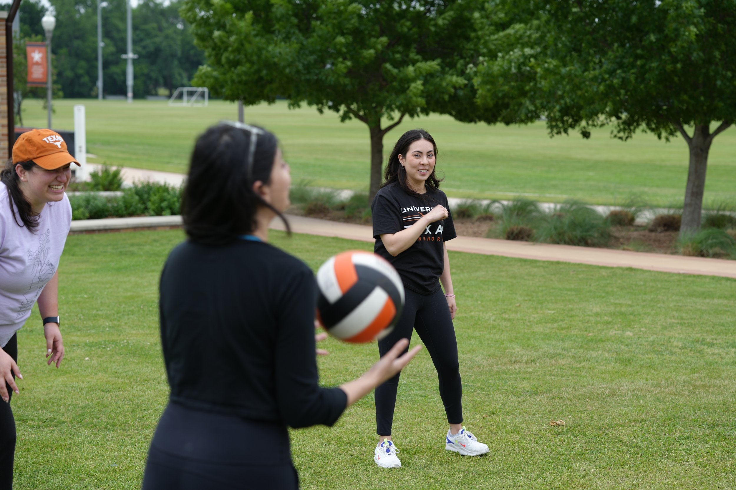 People playing volleyball on a grassy field with trees, a pathway, and a park in the background.
