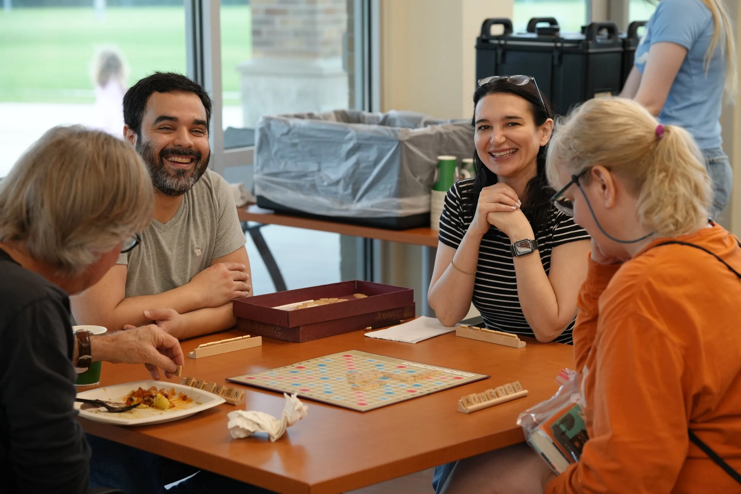 People playing Scrabble at a table, smiling and engaging in the game, with food and game pieces visible.