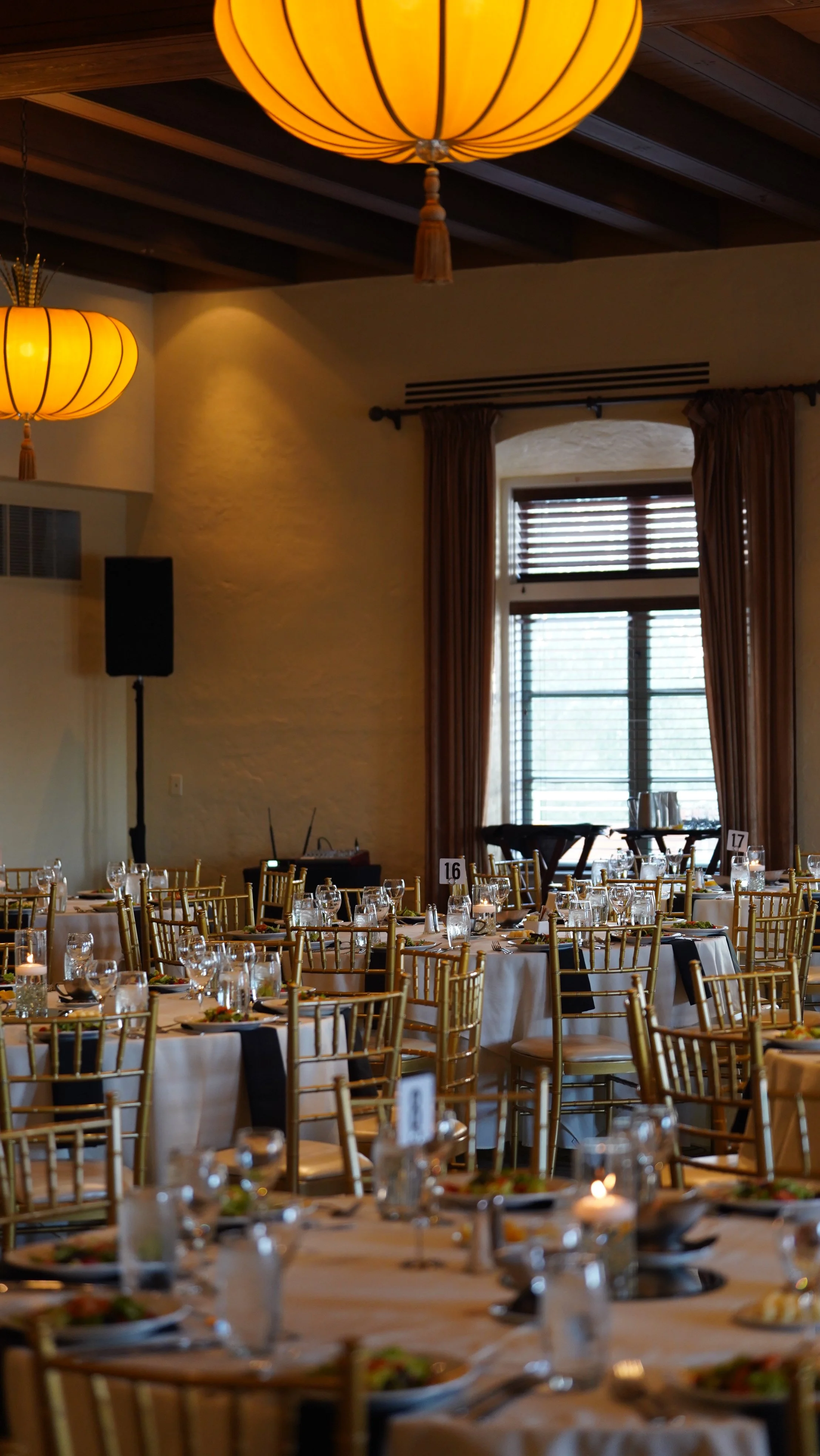 Interior of a banquet hall set up for an event with round tables, gold chairs, tableware, and candle centerpieces, with large windows and warm lighting from hanging lanterns.