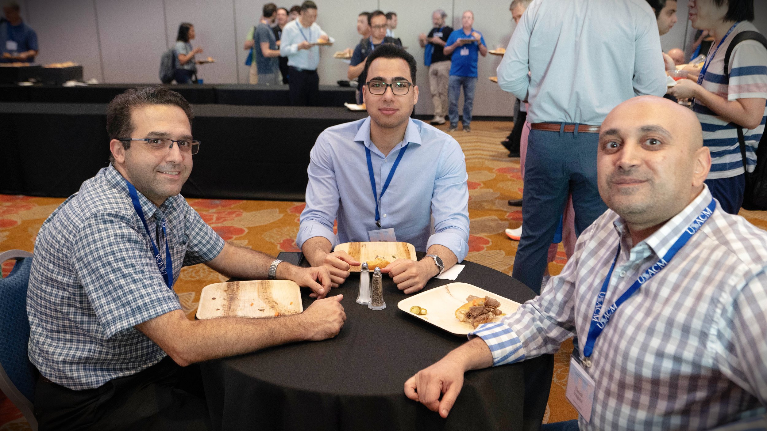 Three men sitting at a round table with plates of food, one holding a wooden plate, in a conference room with other people in the background.