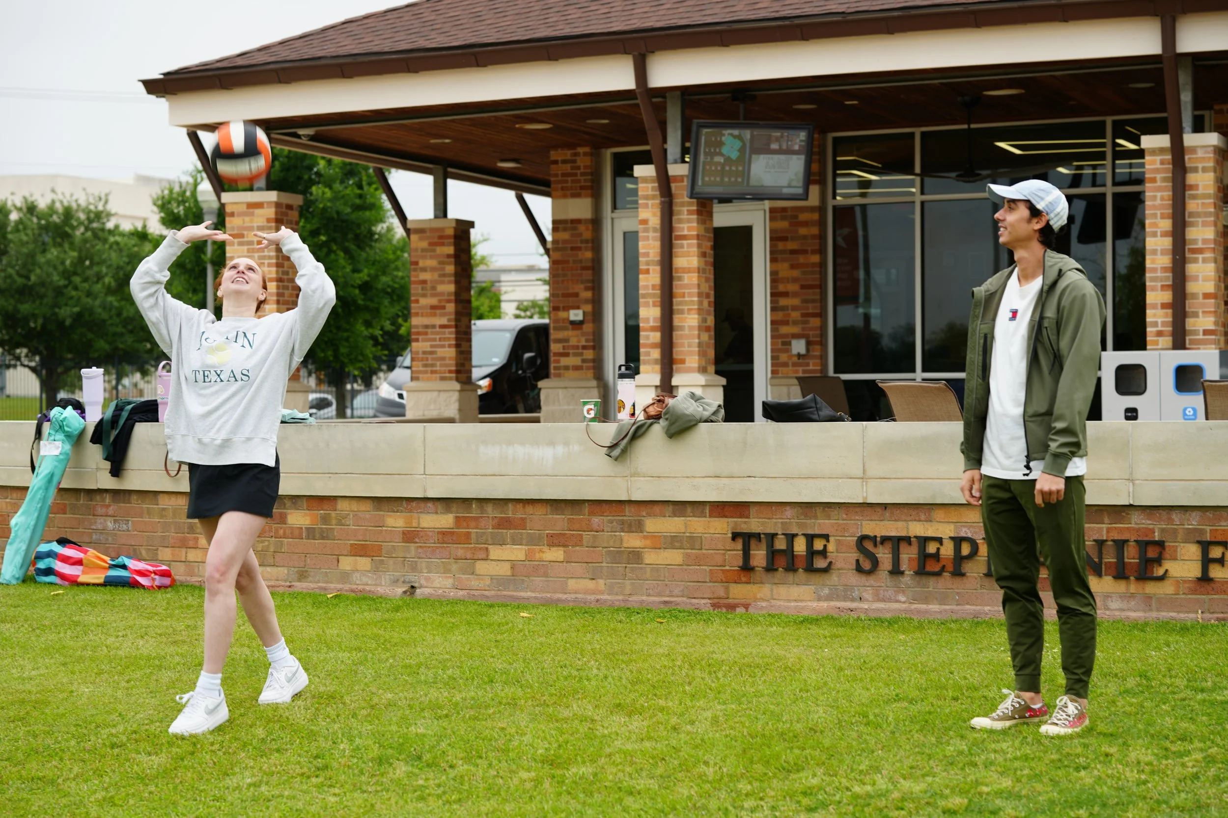 A girl playing with a volleyball outdoors near a brick building, with a young man watching.