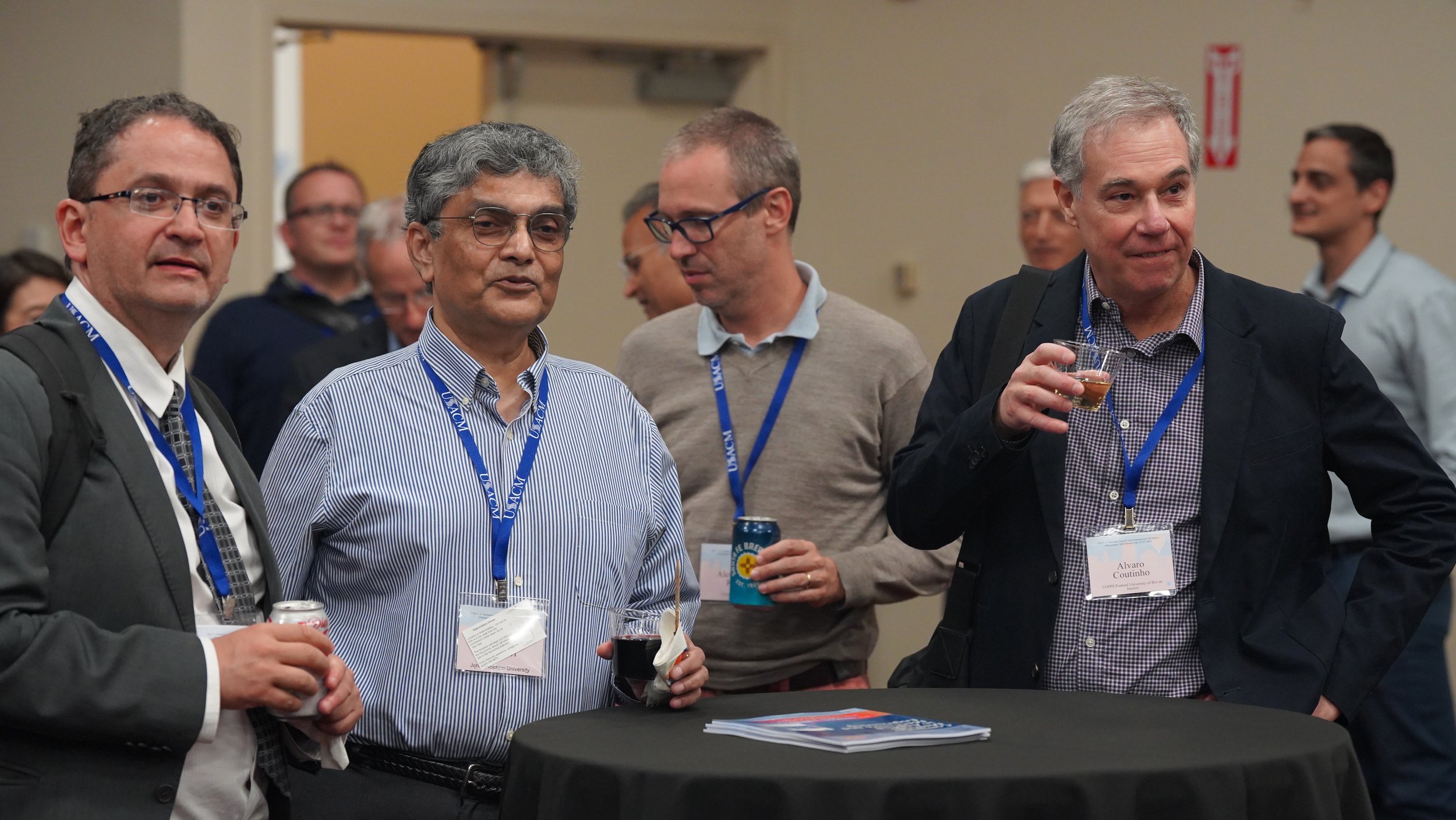 Group of professionally dressed men at a conference or social event, wearing name tags and holding drinks, engaged in conversation.