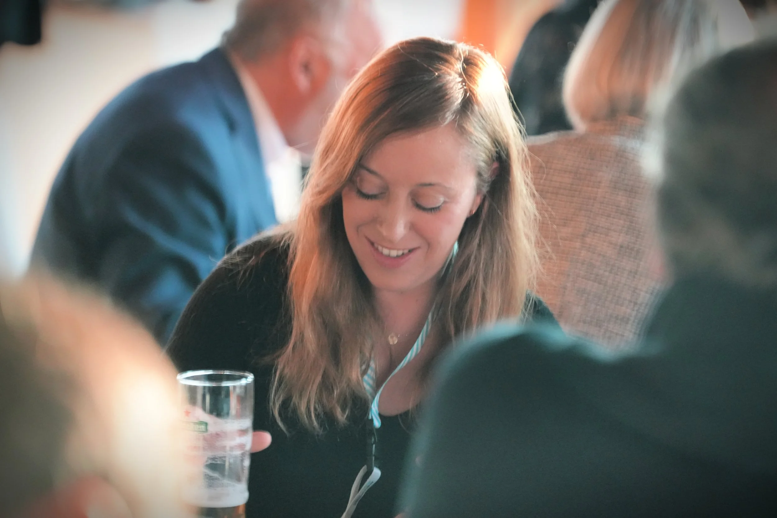 A woman with long brown hair smiling at a social gathering, with blurred people in the background and a glass of water in front of her.