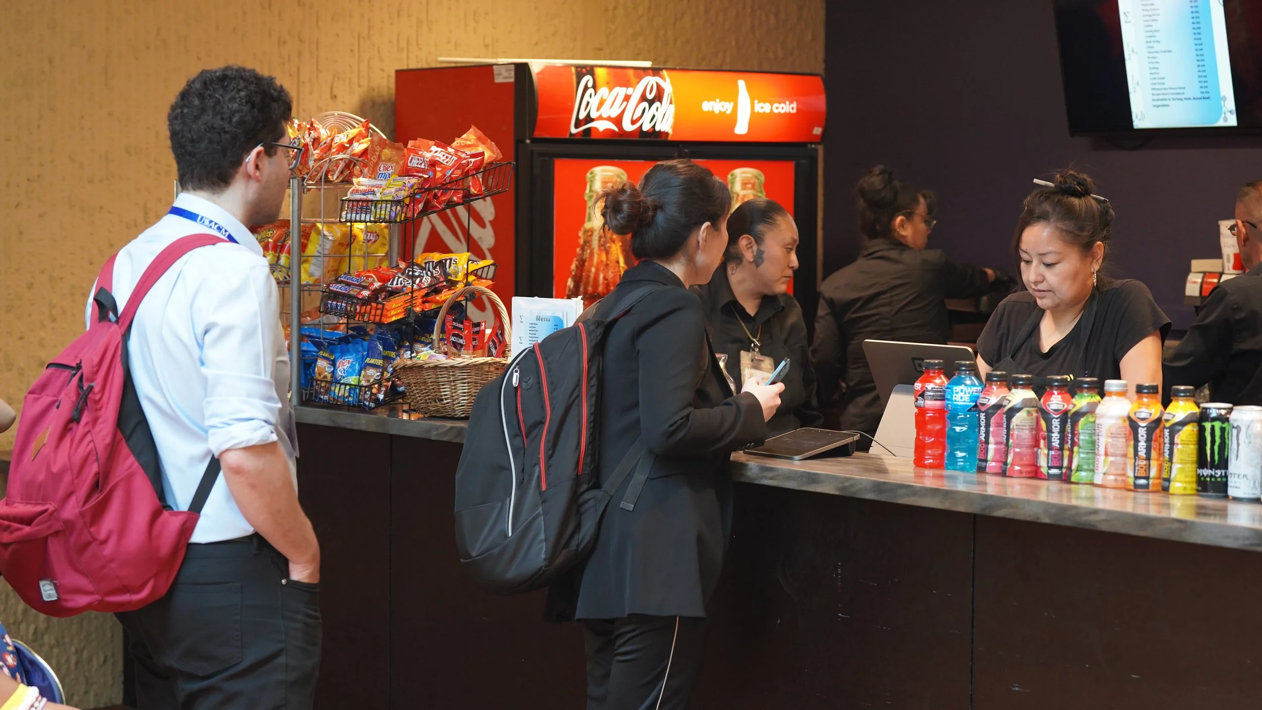 People at a concession counter with snacks, drinks, and a Coca-Cola vending machine in the background.
