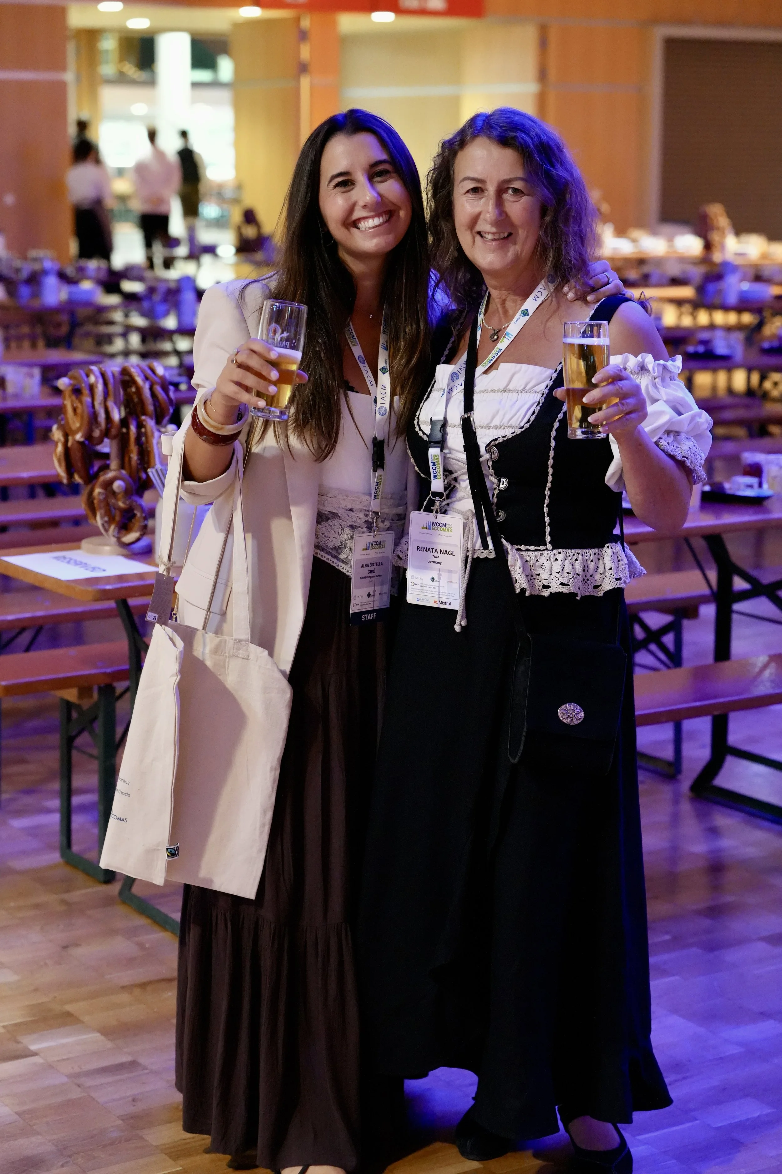 Two women standing together at an indoor event, smiling and holding drinks, wearing event tags. One woman has long dark hair and a white blazer, the other has curly hair and a traditional dress. Background shows other people and event tables.