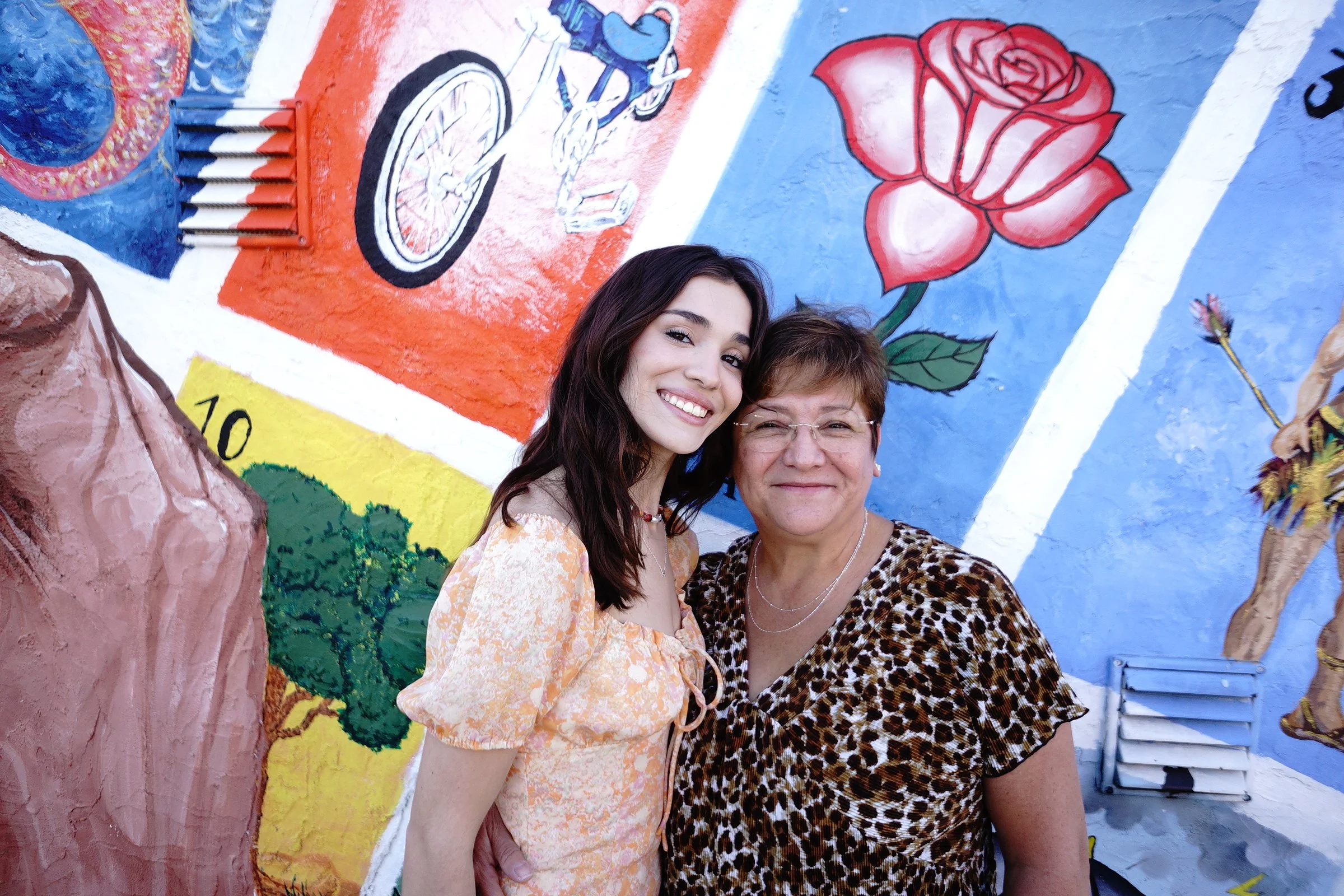 Two women standing in front of a colorful mural featuring a large rose, a bicycle, and abstract designs.