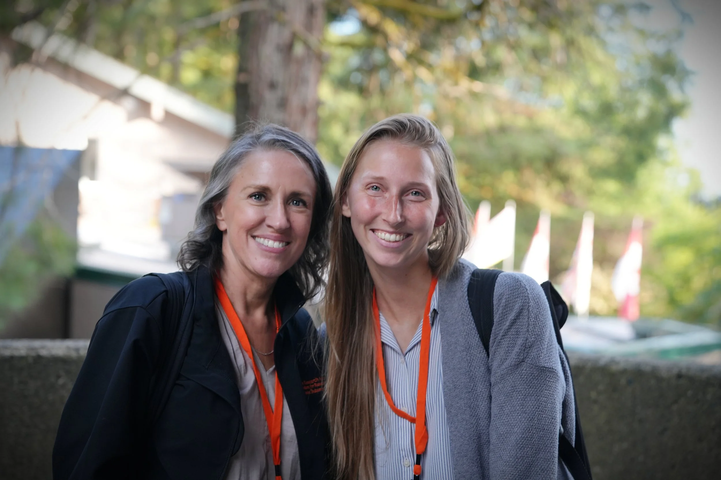 Two smiling women standing outdoors with trees and flags in the background, wearing casual clothes and orange lanyards.