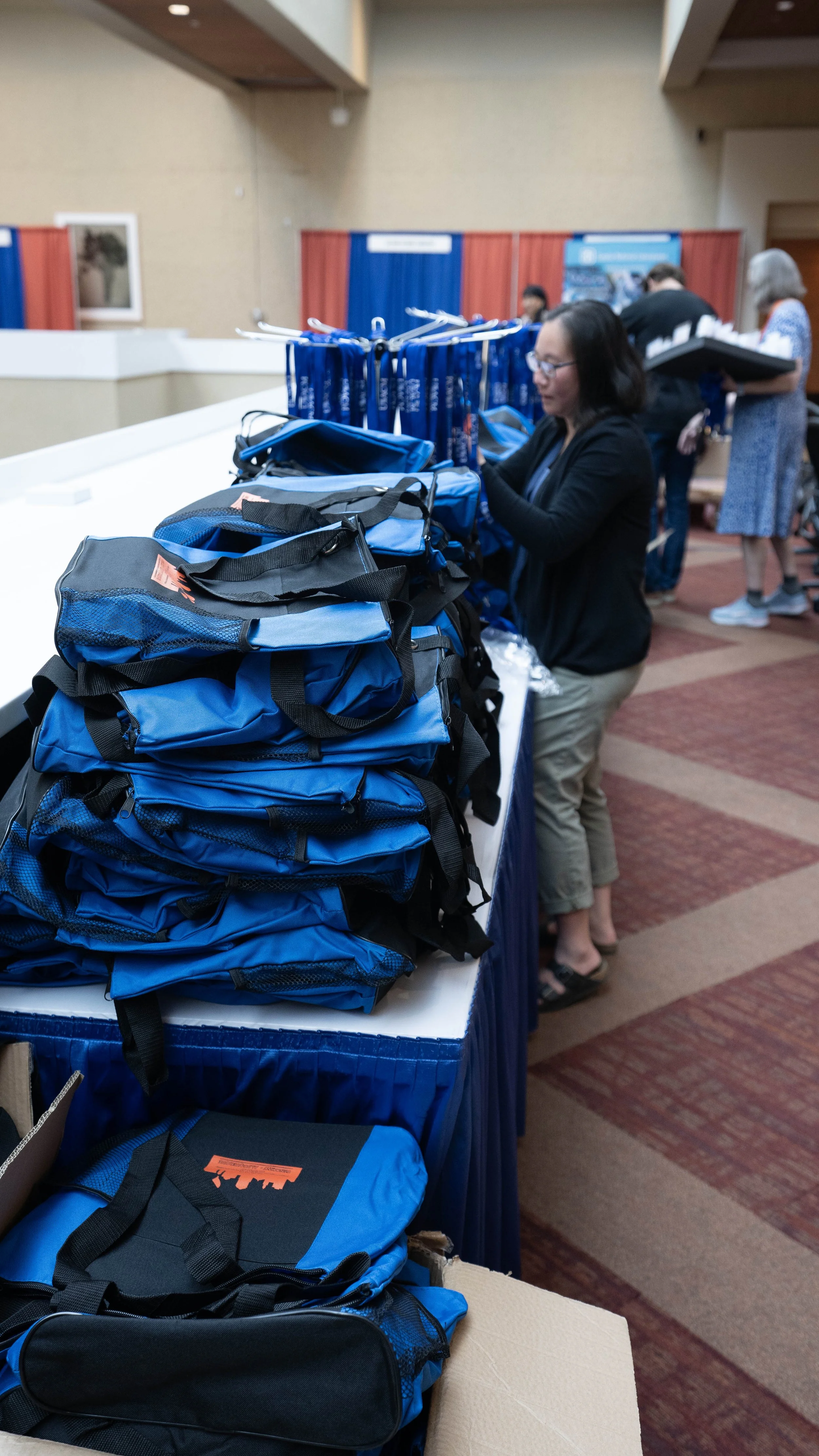 Table with blue backpacks and bags at a conference or event. People in the background browsing the display.