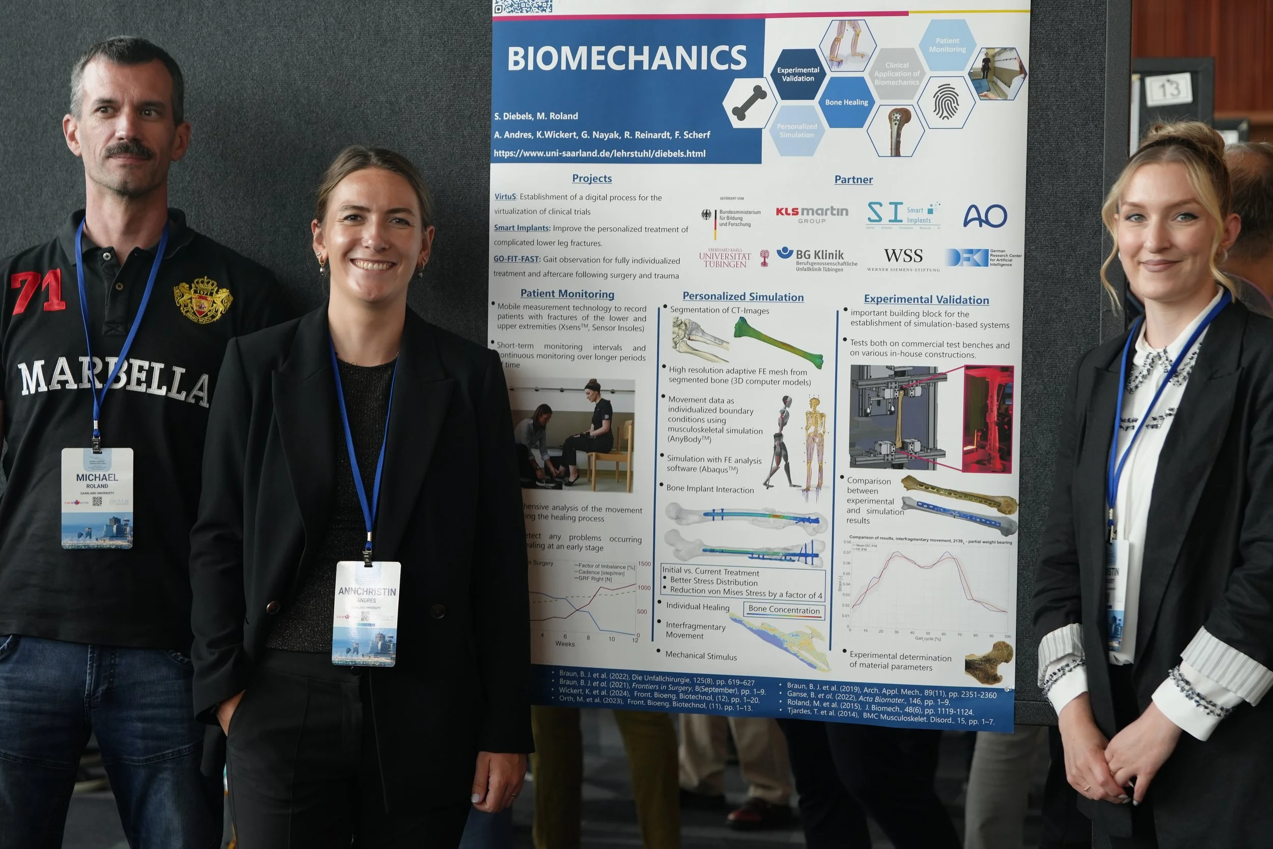Three people standing next to a scientific research poster at an academic conference. The poster is titled "Biomechanics" and features diagrams and text about research projects. Two women and one man are smiling and wearing conference badges.