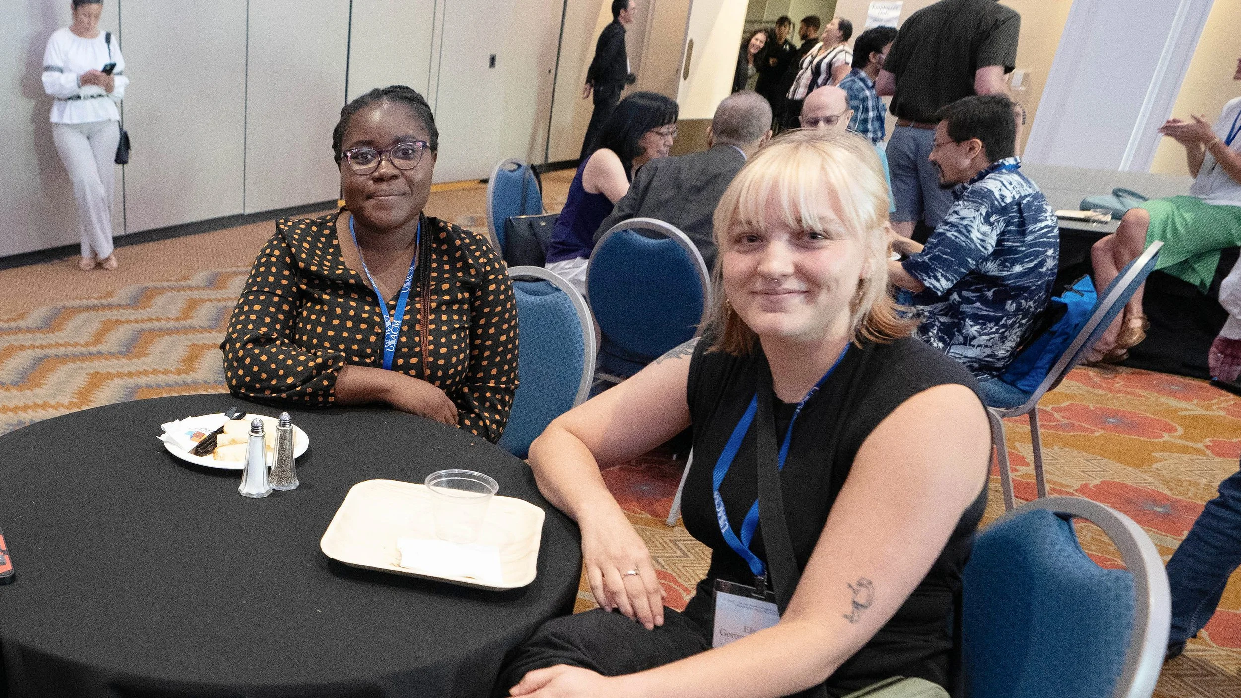 Two women are seated at a round table in a conference or event setting, smiling at the camera. One woman has dark skin and glasses, wearing a black dress with orange polka dots. The other woman has light skin, blonde hair, and is wearing a black sleeveless top. On the table are some condiments, a plate with some small food items, and a napkin. In the background, other attendees are seated or walking around, and a woman is standing against a wall looking at her phone.