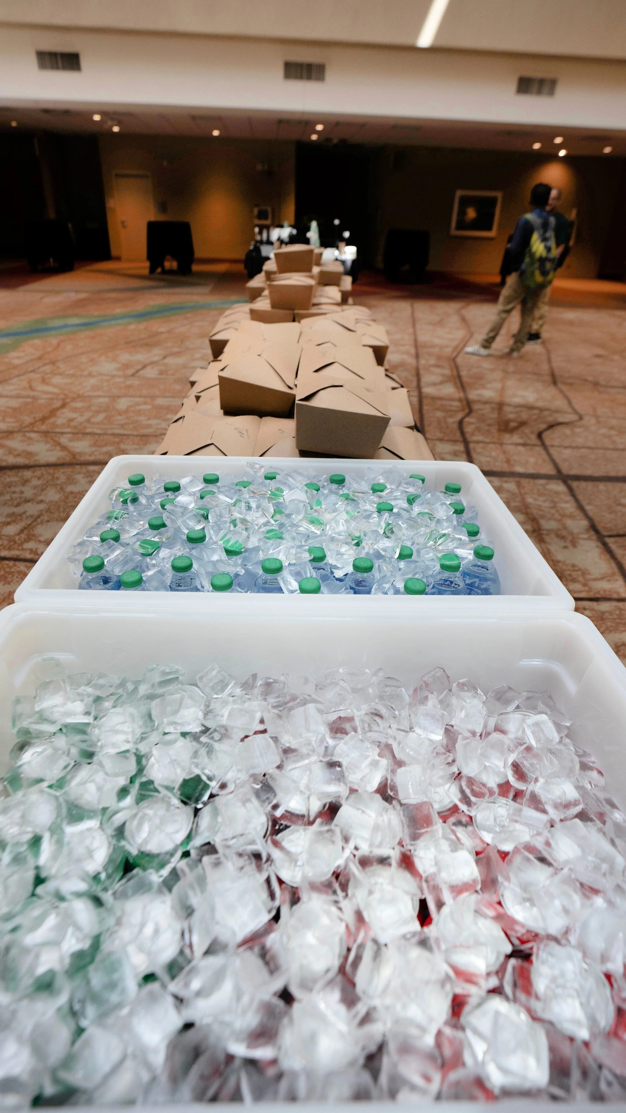 A table with bottled water, paper food containers, and ice in a conference or event hall.