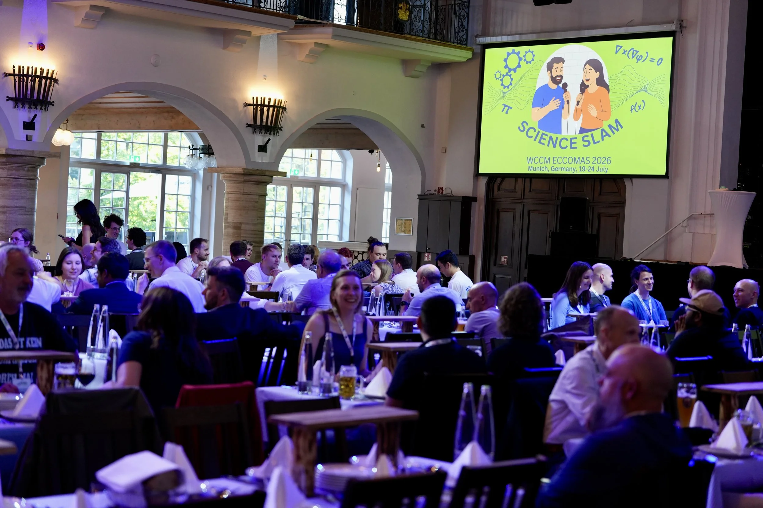 A large indoor conference or event hall filled with many people seated at tables, with some standing and talking. There is a big screen at the front displaying a logo for 'Science Slam' with two cartoon people holding microphones, and text indicating the event is part of WCCM ECCOMAS 2026 in Munich, Germany, from July 19 to 24. The room has bright windows, arched architecture, and decorative wall sconces.