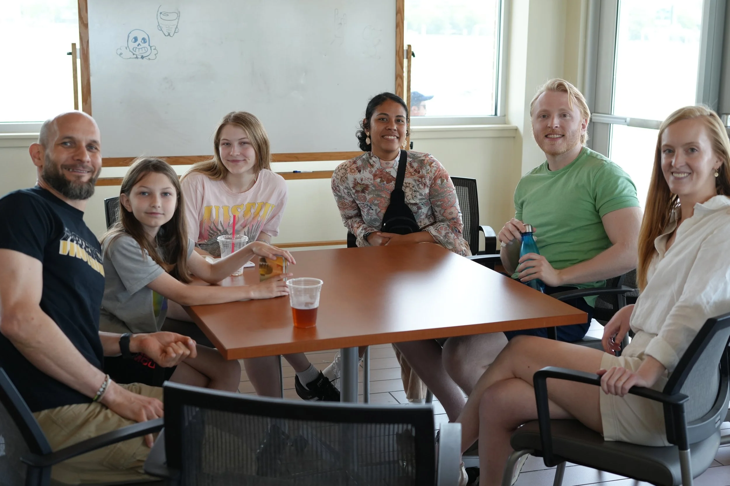 Group of six people sitting around a conference table in a bright room with large windows. They are smiling and engaging in conversation. There are drinks on the table.