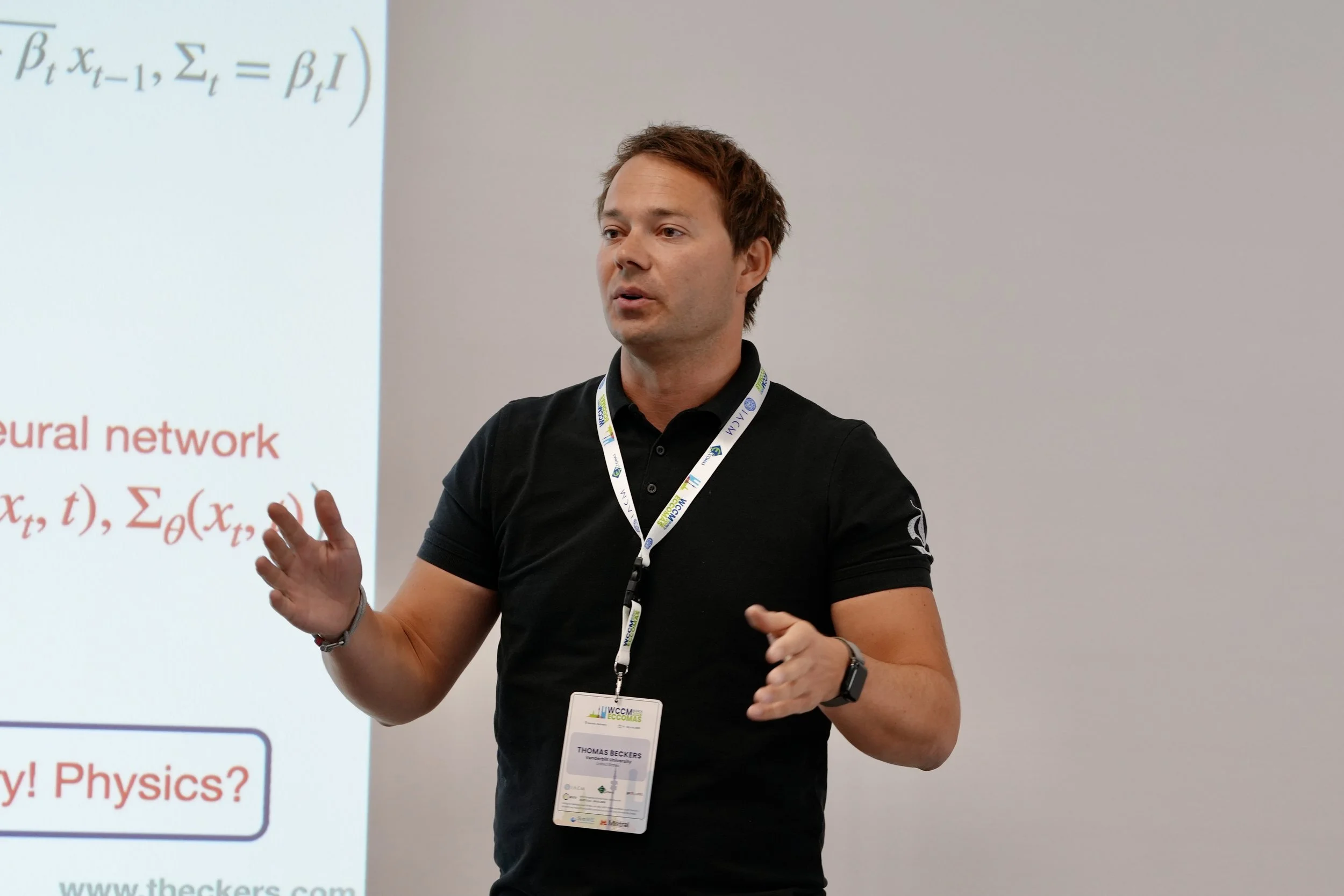 A man with short brown hair wearing a black polo shirt and a conference badge on a lanyard is giving a presentation. Behind him, there's a large screen displaying mathematical equations and text related to neural networks and physics, with some red-colored words.