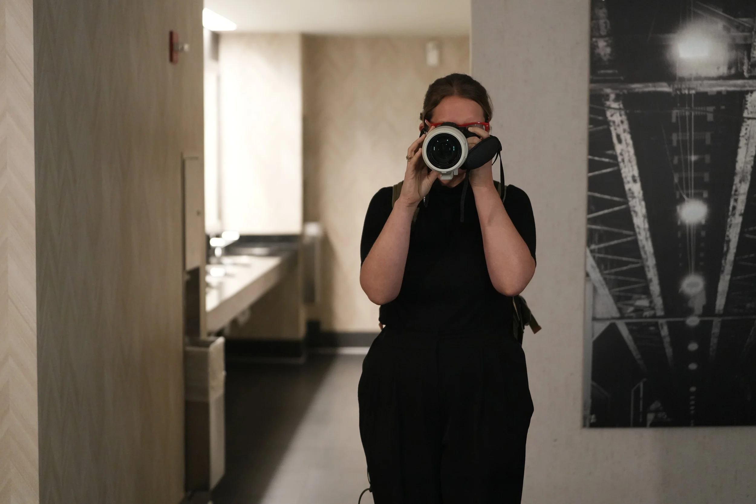 Person with dark hair in a black shirt taking a photo with a camera in a hallway, behind a mirror, with artwork on the wall.