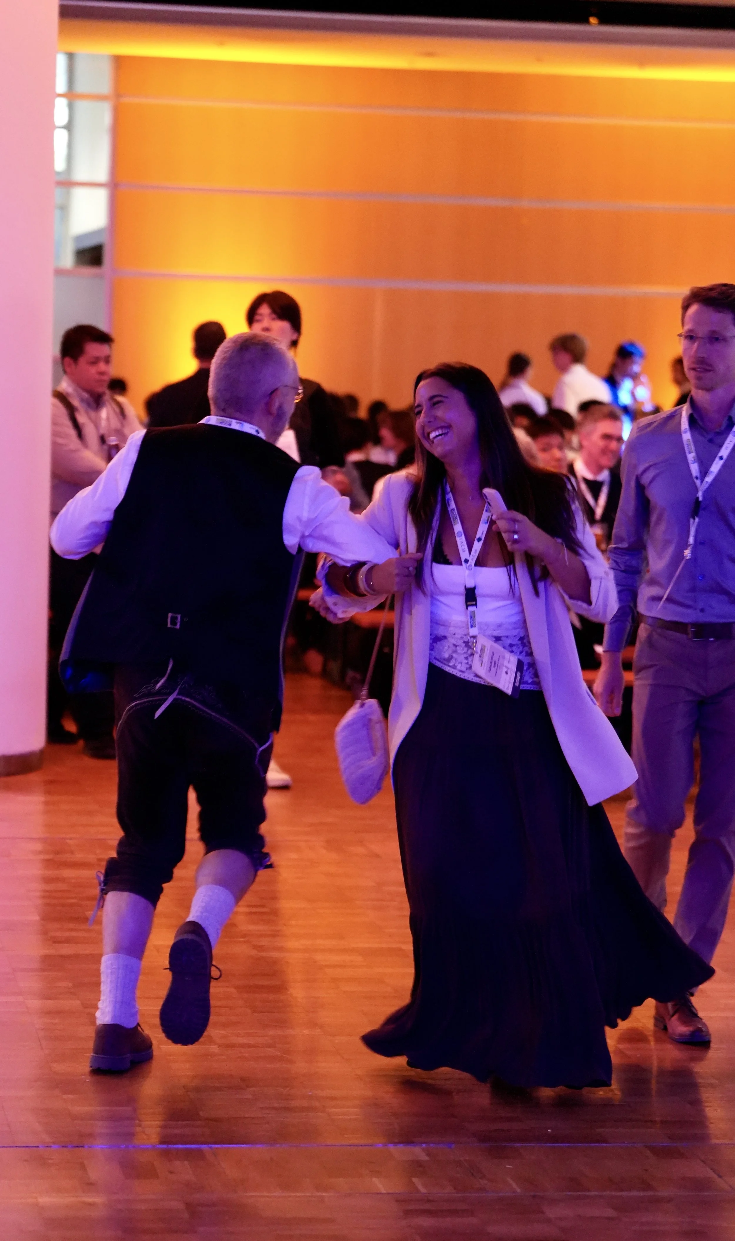 A group of people dancing indoors at a conference or event, with vibrant orange lighting on the wall behind them.