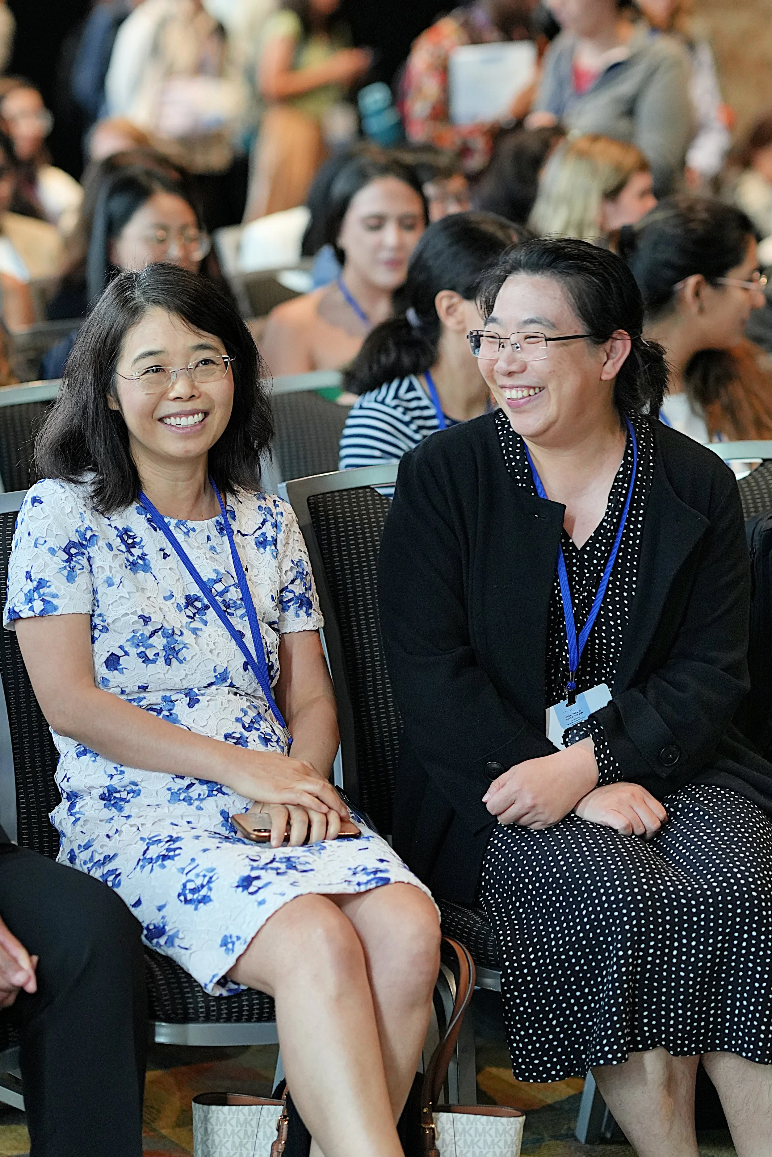 Two women with glasses smiling and engaging in conversation while sitting among a large group of people at an indoor event or conference.