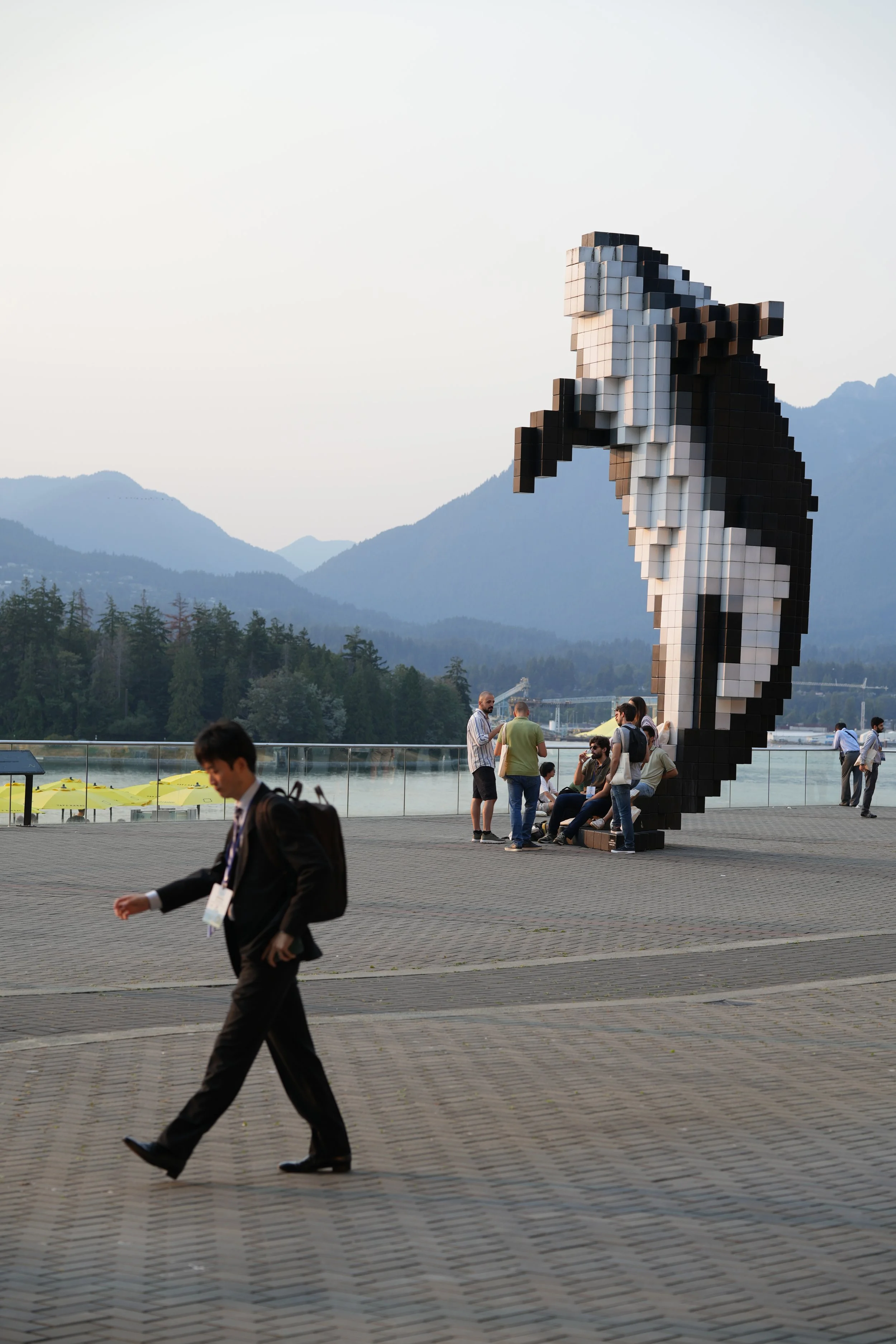 People gathered around a large pixelated sculpture of a horse's head outdoors with mountains in the background and a lake.