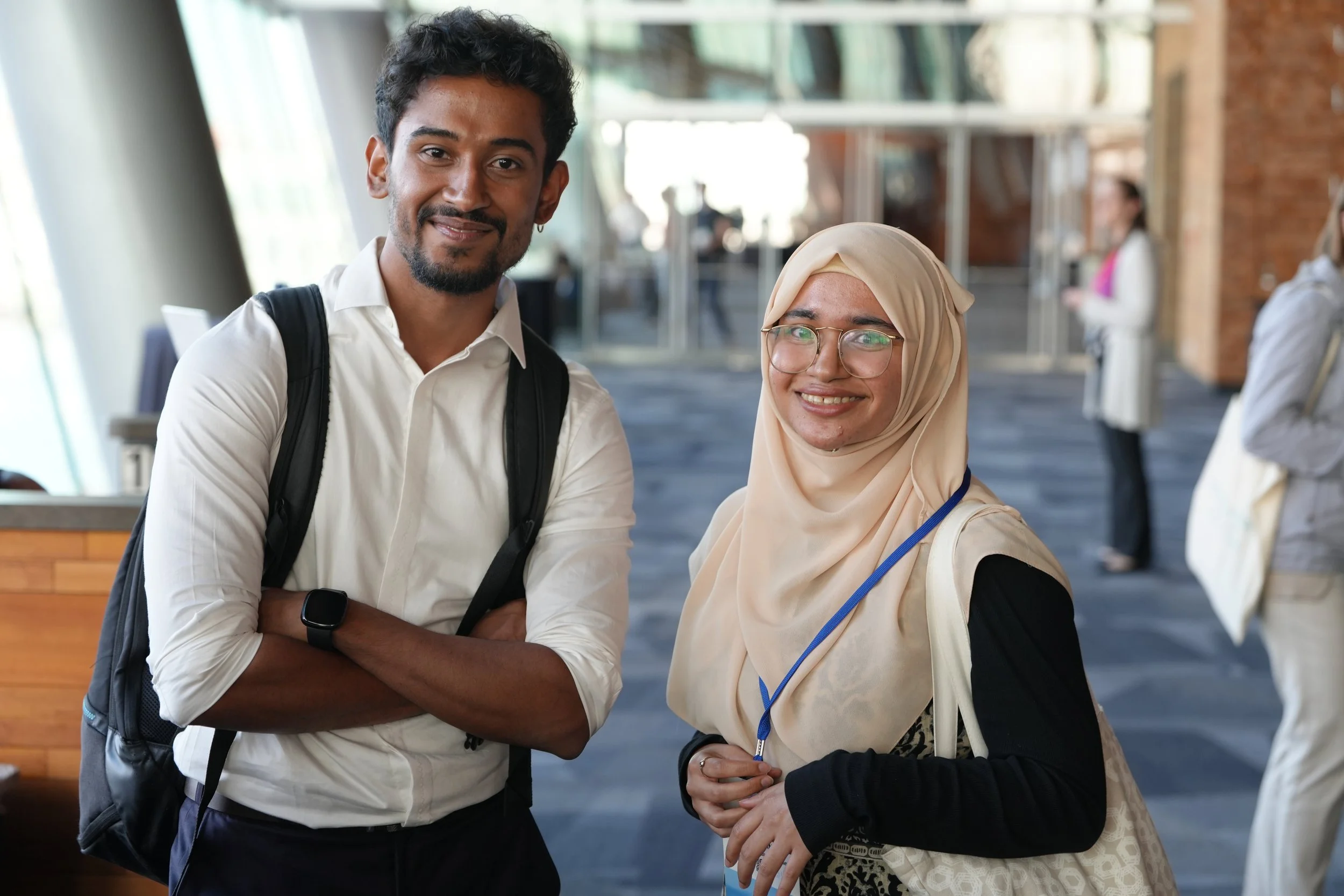 A man with dark hair, a beard, wearing a white shirt and a smartwatch, standing with his arms crossed, smiling at the camera. Next to him is a woman wearing glasses and a beige hijab, also smiling. The background shows a modern, open indoor space with large windows, brick walls, and other people in the distance.