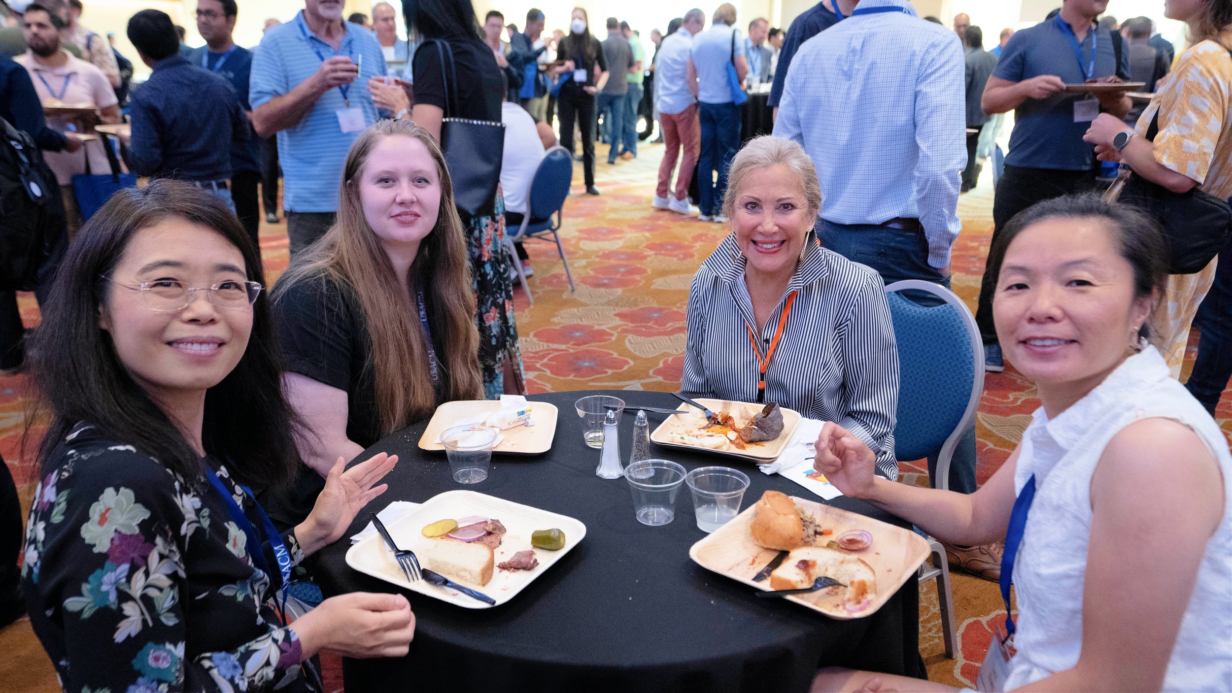 Four women sitting at a round table with lunch trays, smiling, in a busy conference or event setting.