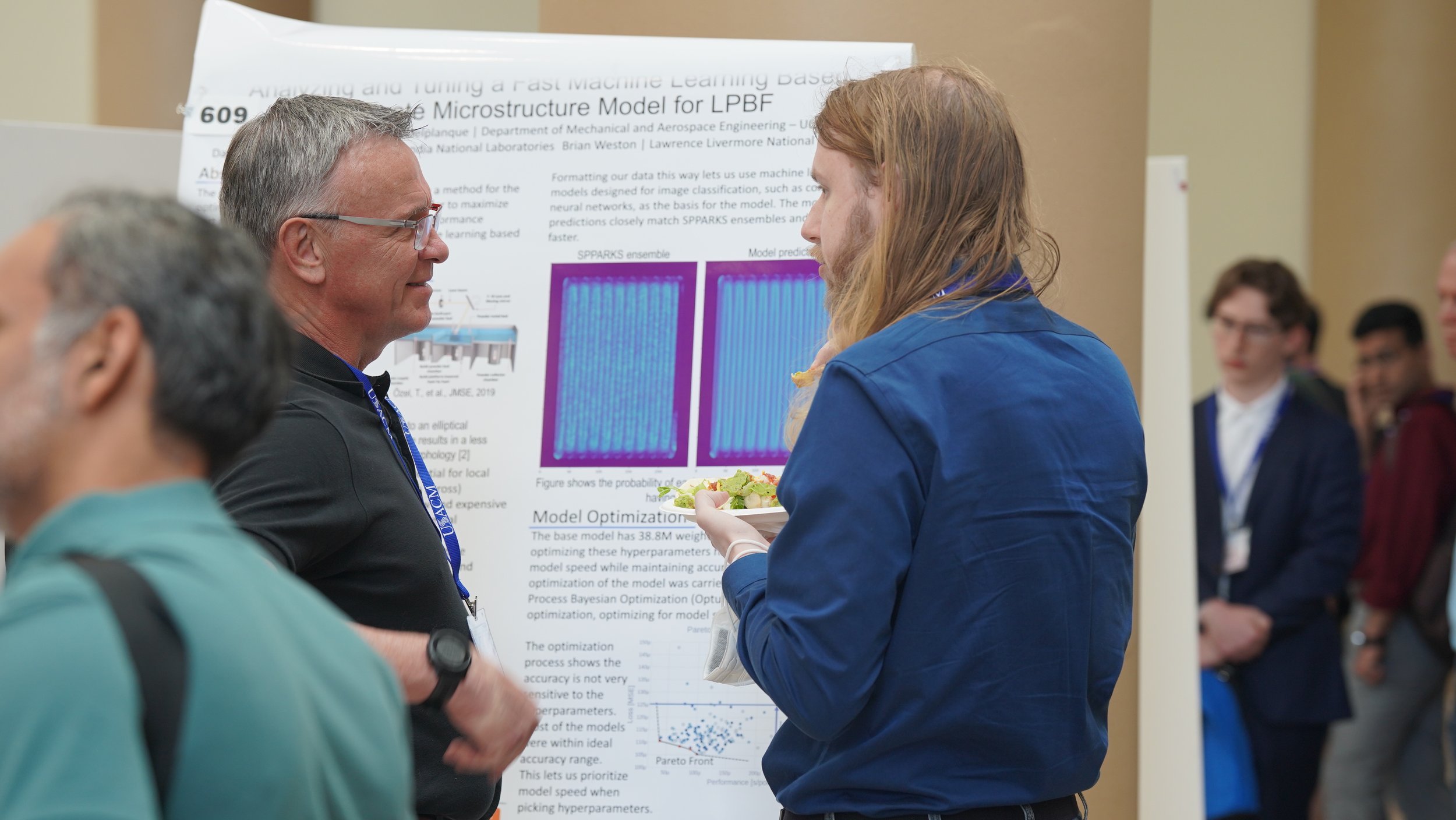A young woman in a blue shirt holding a plate of food is conversing with a gray-haired man wearing glasses and a dark shirt at a research poster presentation. The poster displays scientific data, charts, and text on microstructure models.