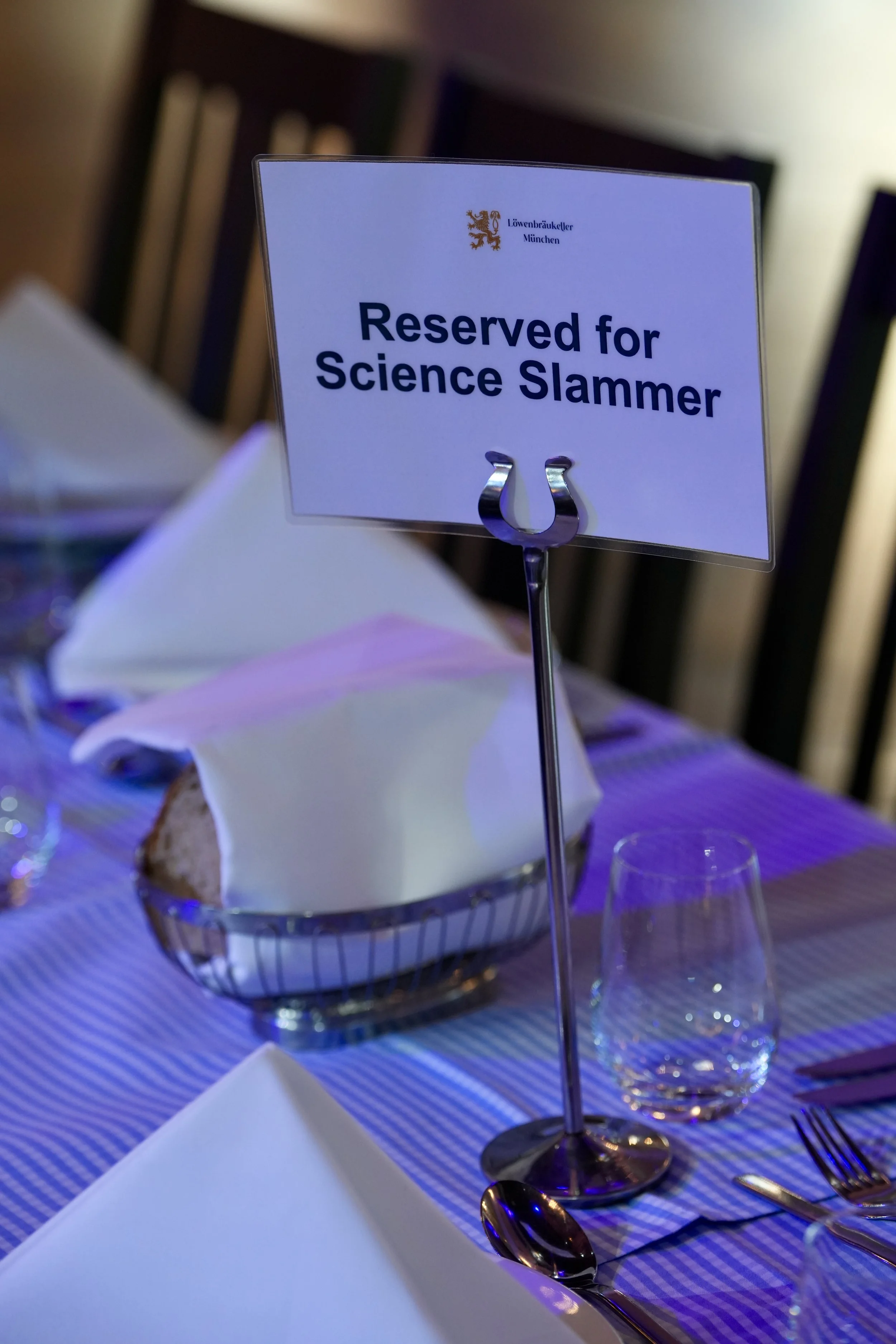 A table setup at a formal event with a sign reading 'Reserved for Science Slammer.' The table has a glass of water, a white napkin, silverware, and a cake in a basket.
