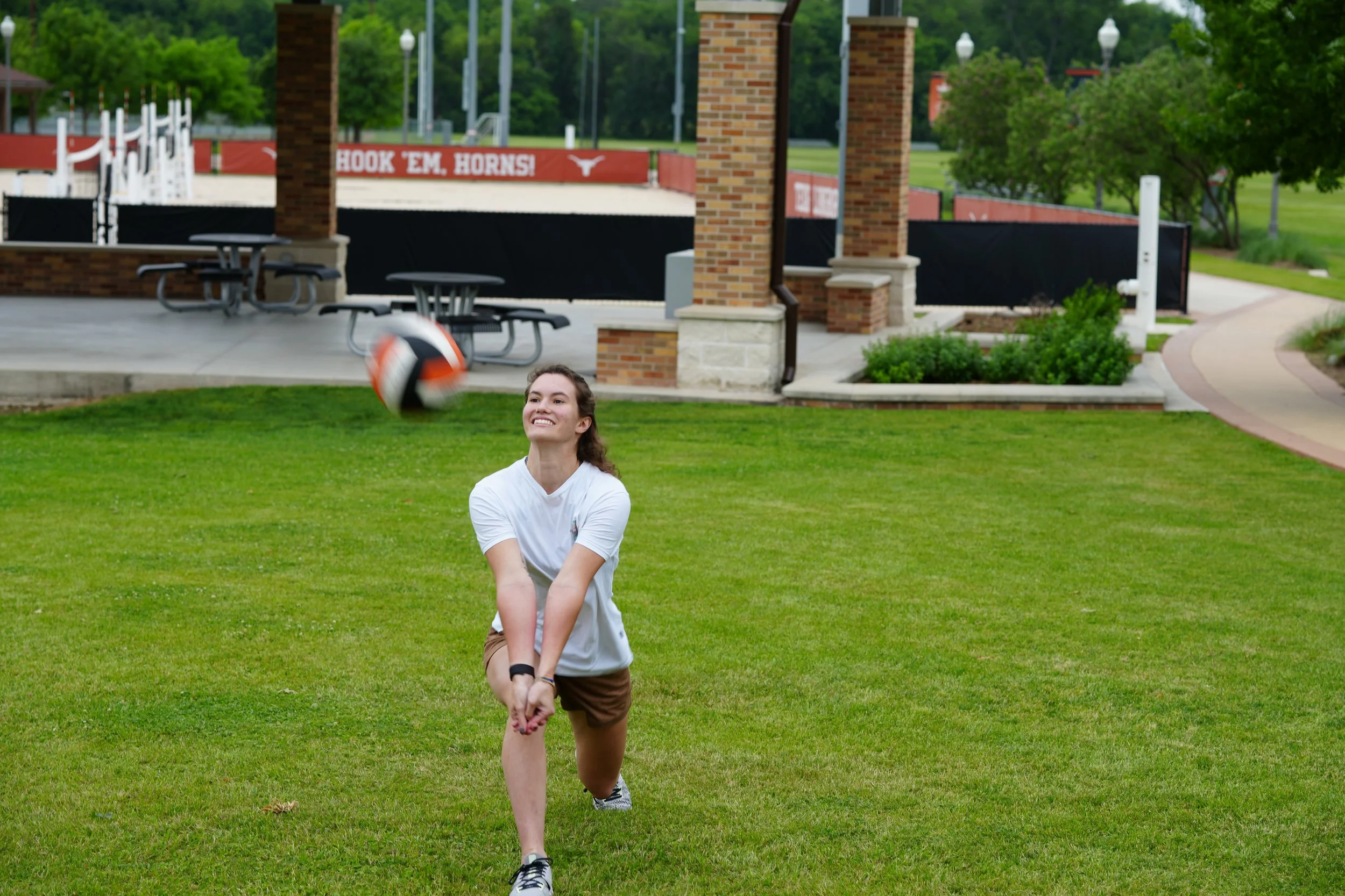 A young girl in a white shirt and brown shorts is playing with a volleyball, preparing to hit it. She is outdoors on a grassy area near a park or sports field, with a brick building, benches, and trees in the background.