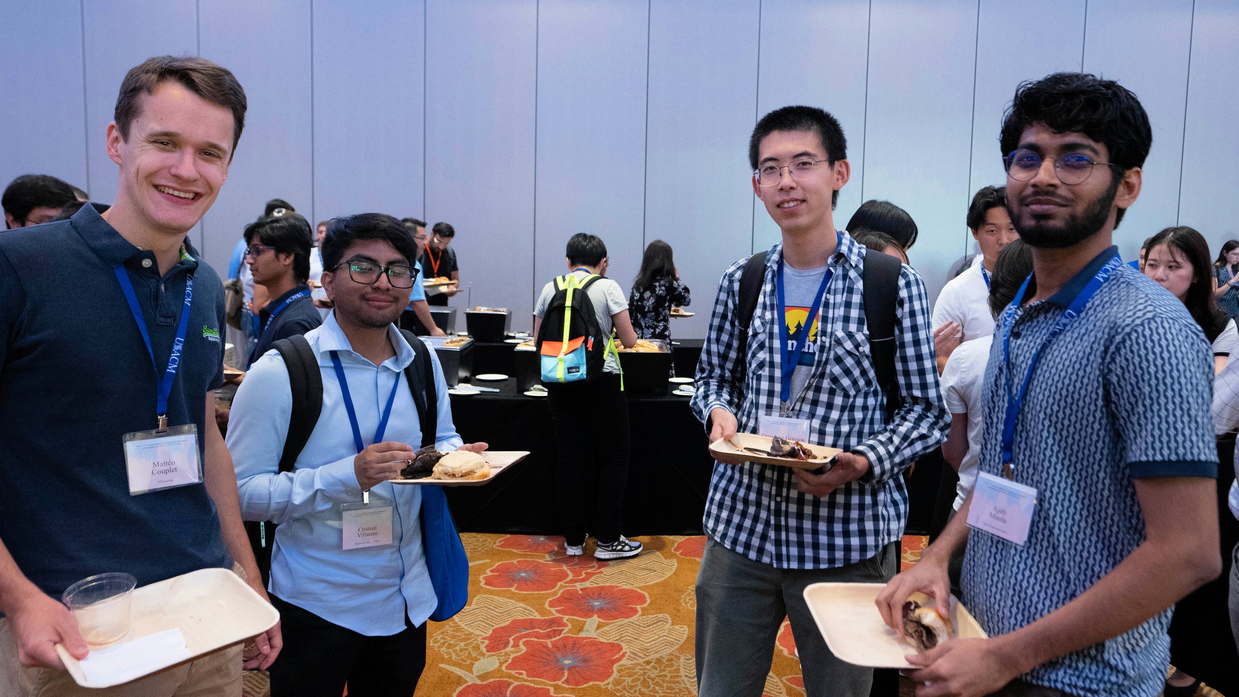 Group of four young men standing together at a conference, holding plates of food, with other attendees and a buffet table in the background.