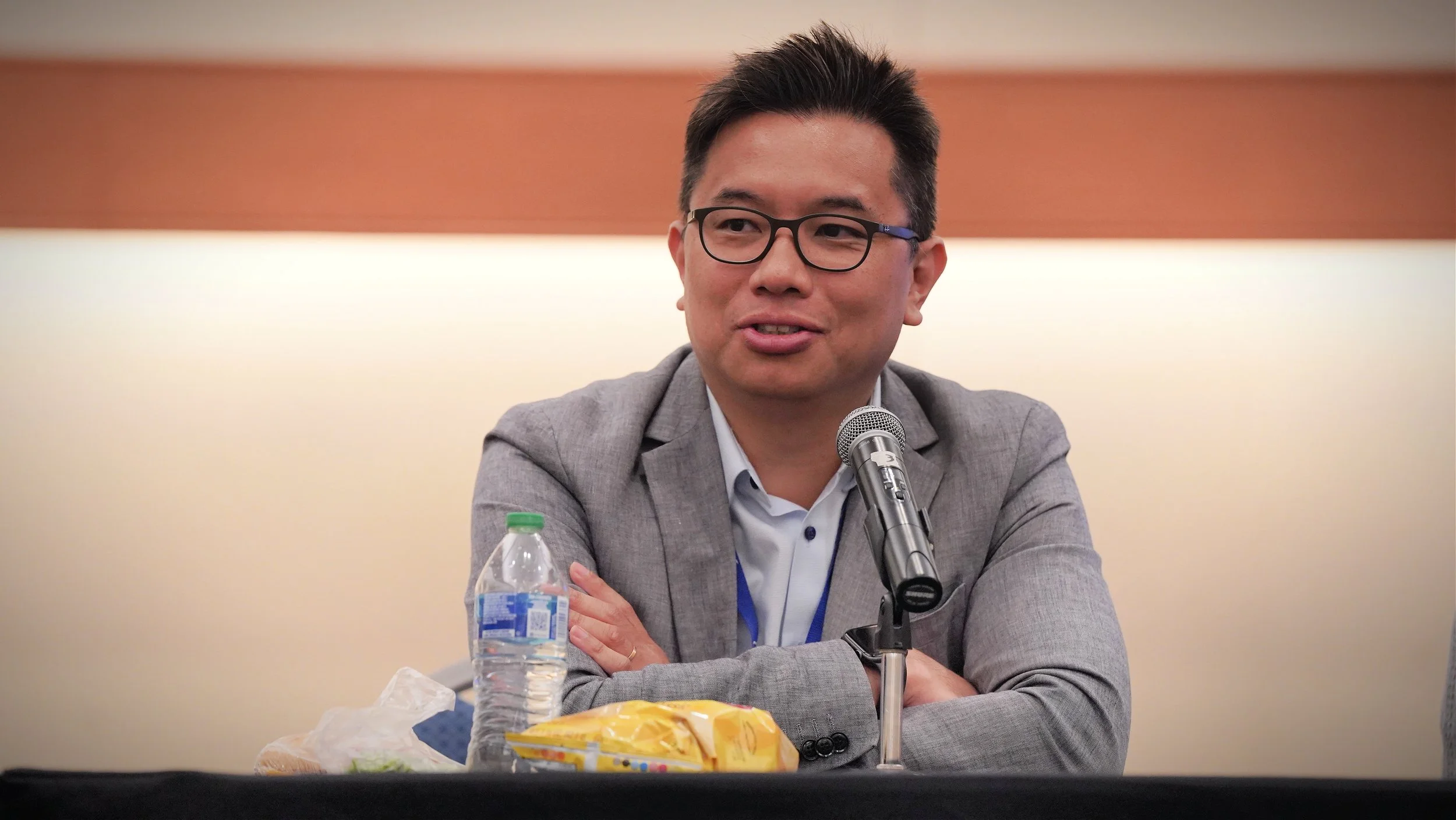 A man wearing glasses and a gray suit sitting at a table with a microphone in front of him, a water bottle, snacks, and a blue lanyard, speaking at a conference or event.