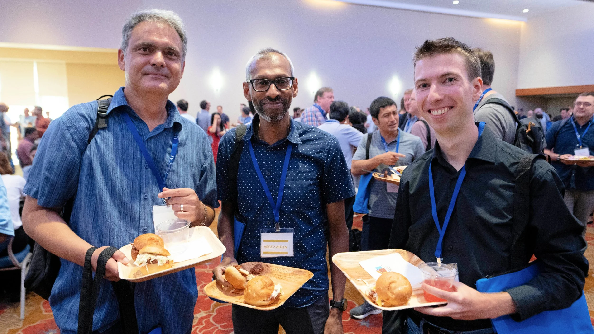 Three men at a conference holding trays with vegan sliders and drinks. They are smiling and wearing conference badges, and there is a crowd in the background.