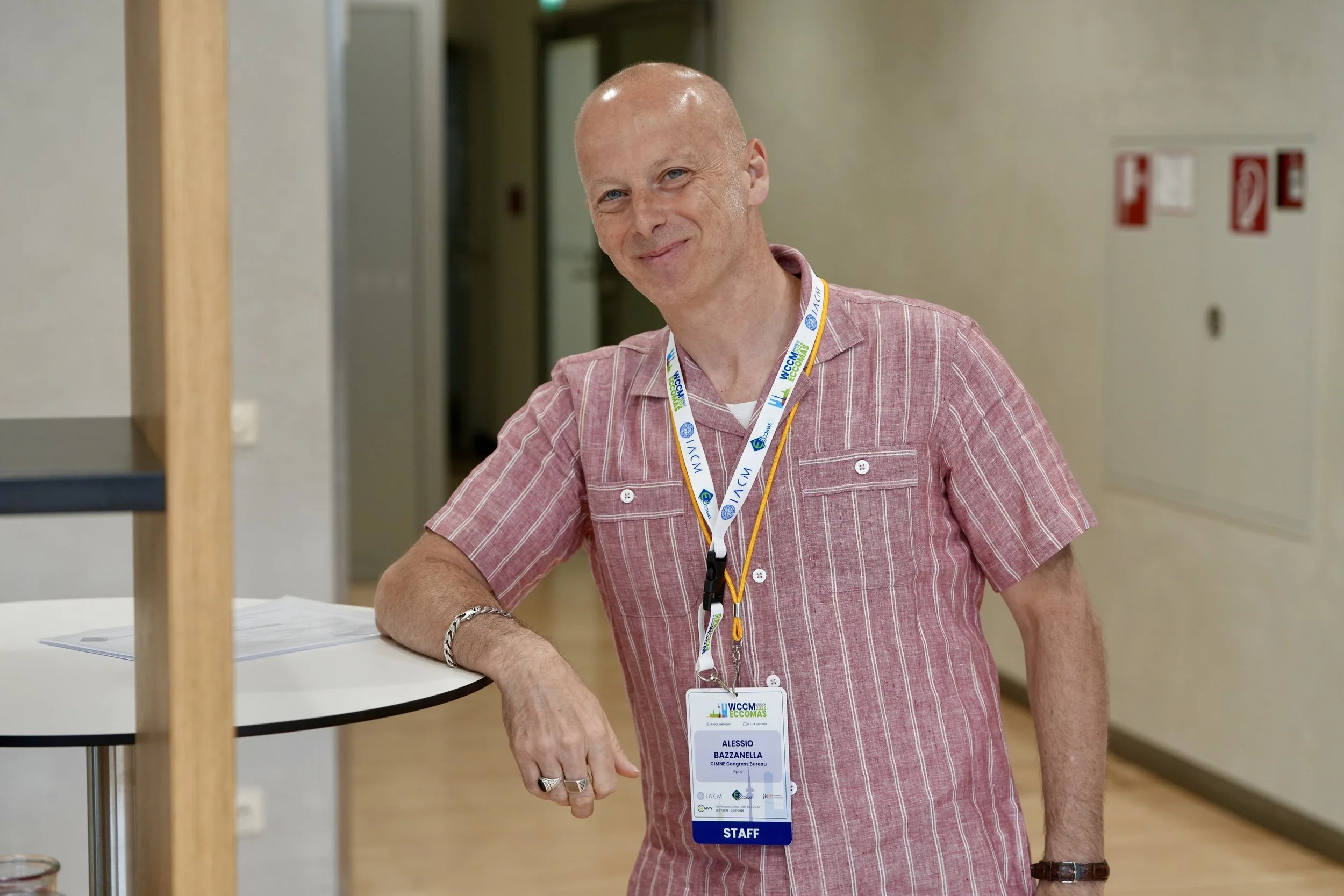 A man with a bald head and blue eyes, wearing a pink striped shirt and a conference badge, smiling and standing next to a table in a conference hallway.