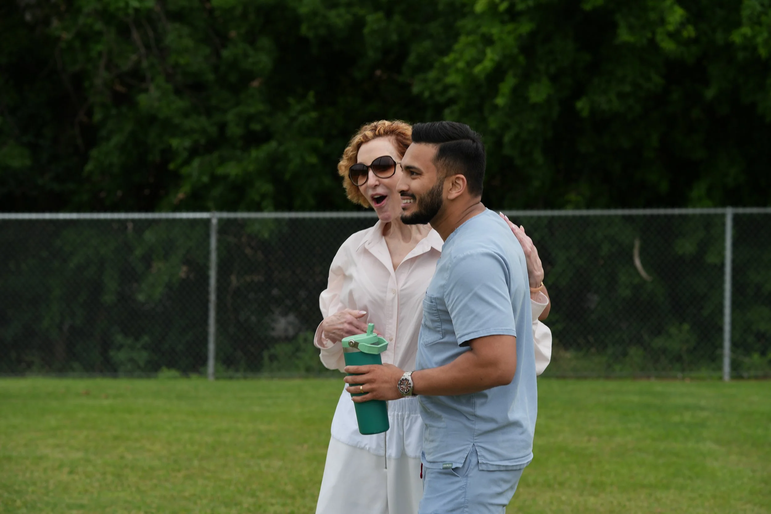A woman with curly red hair and sunglasses speaking to a man with dark hair and beard, both smiling, standing on a grassy field near a chain-link fence with green trees in the background.