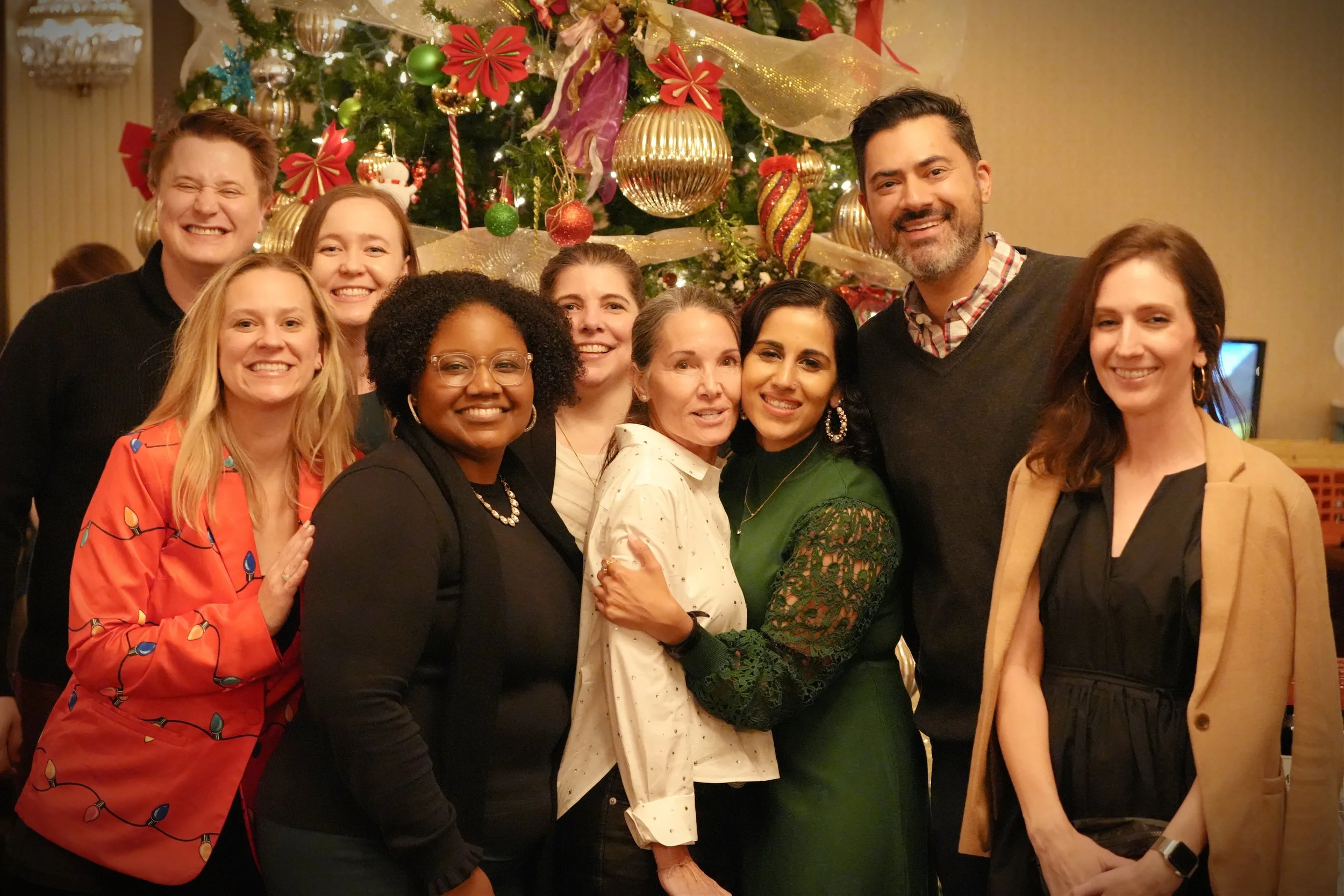 A group of ten diverse people posing in front of a decorated Christmas tree, smiling and celebrating together at a holiday gathering.