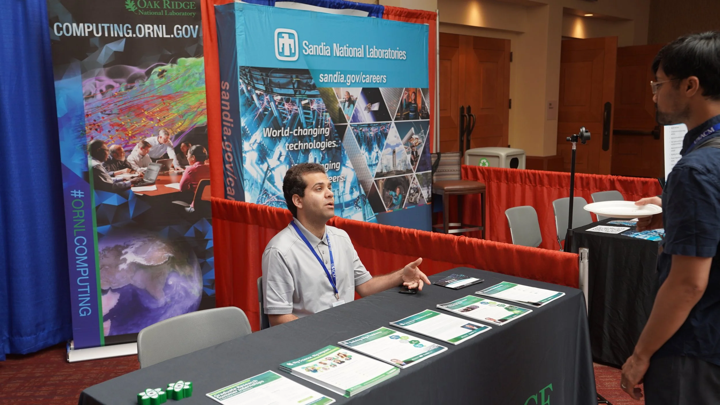 A man with dark hair and a goatee, wearing glasses and a dark shirt, is standing and talking to a seated man with short dark curly hair, wearing a light gray polo shirt, at a booth in a conference or expo. The booth has a large blue and red banner for Sandia National Laboratories with images related to science and technology. There are brochures on the table and around the booth, and the background shows other conference banners and booths.