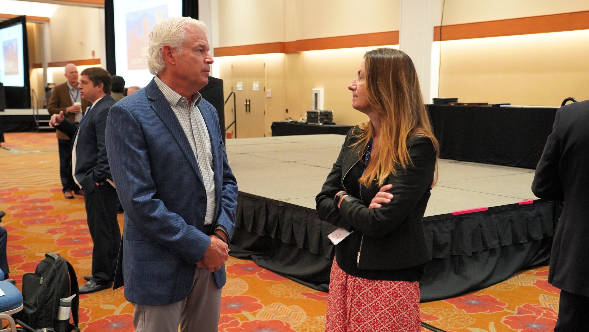 Two people, a man with white hair wearing a blue blazer and a woman with long brown hair wearing a black jacket, are standing face to face and having a conversation at an indoor conference or event.