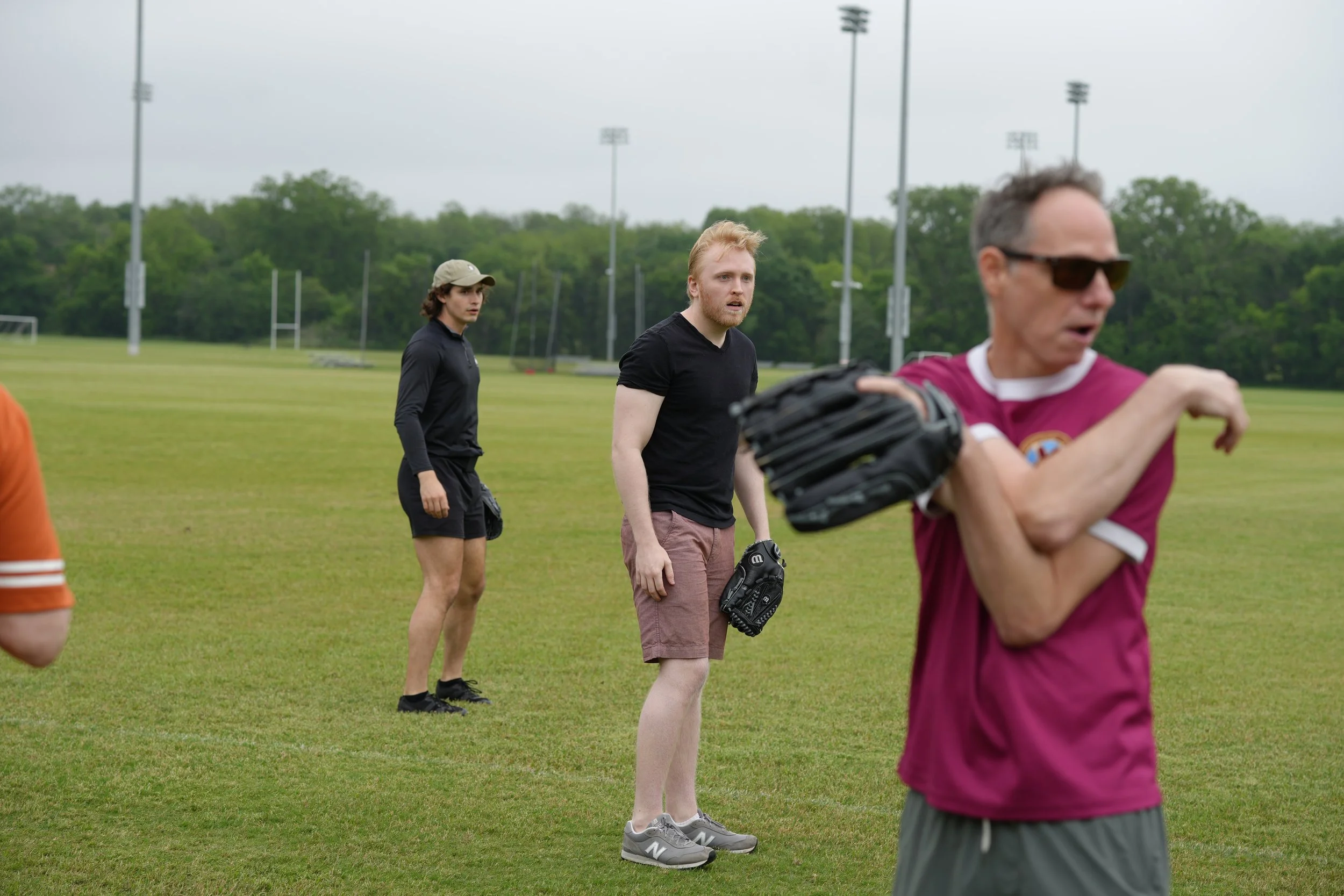 People on a baseball field engaging in a casual activity or conversation, with one person holding a baseball glove, and a man in sunglasses and a maroon shirt in the foreground.