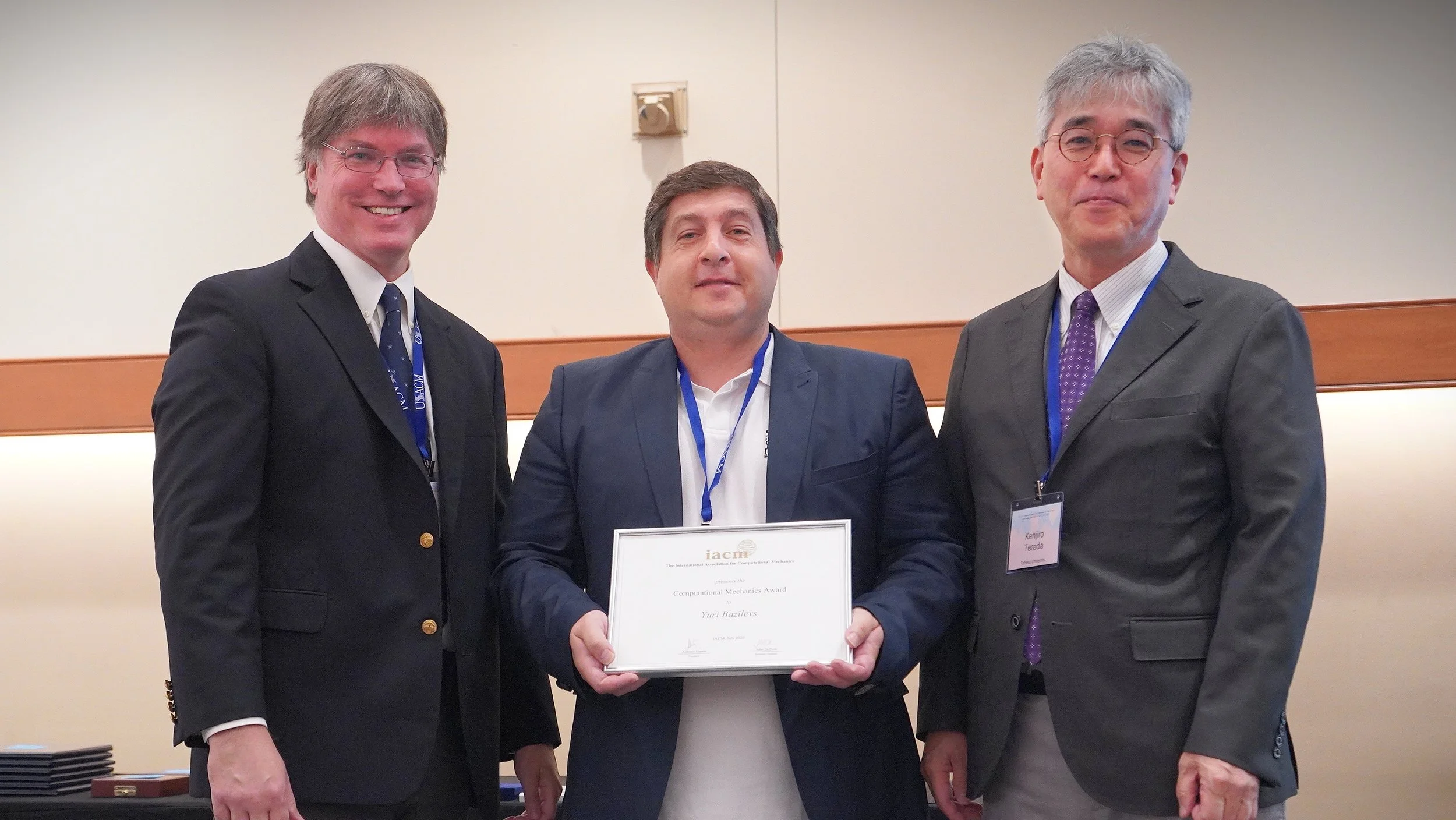 Three men in suits standing together at an award ceremony, with the man in the middle holding a framed certificate.
