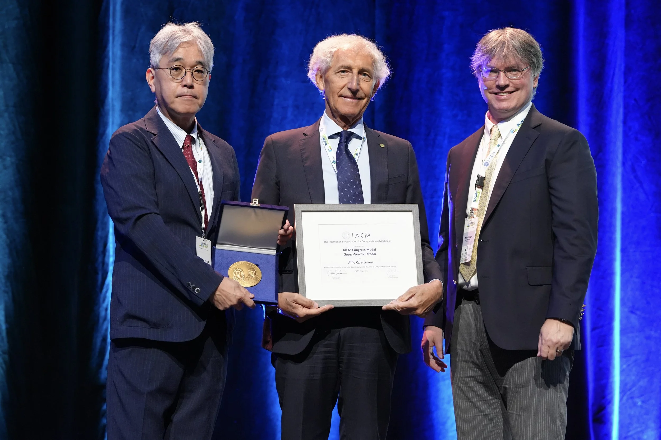 Three men in suits standing on a stage with a blue curtain backdrop. The man on the left is holding a medal and a certificate, while the man in the middle is handing him the certificate. The man on the right is smiling and watching the presentation.