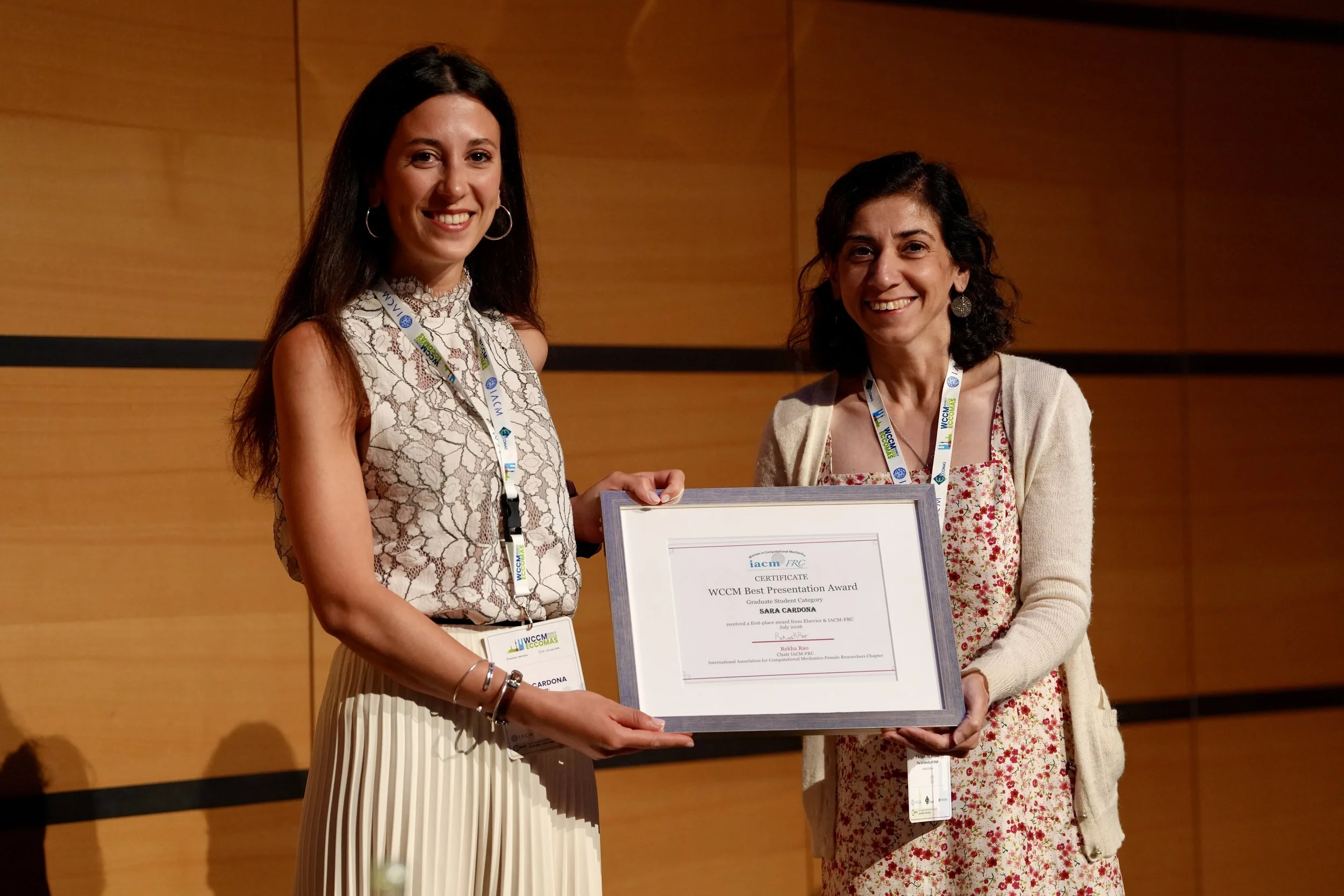 Two women are smiling and holding a large framed certificate during an award ceremony in a conference room with wooden panel walls. One woman is wearing a white dress with a lace pattern, while the other is dressed in a floral dress with a cream cardigan. Both women have conference badges hanging around their necks.