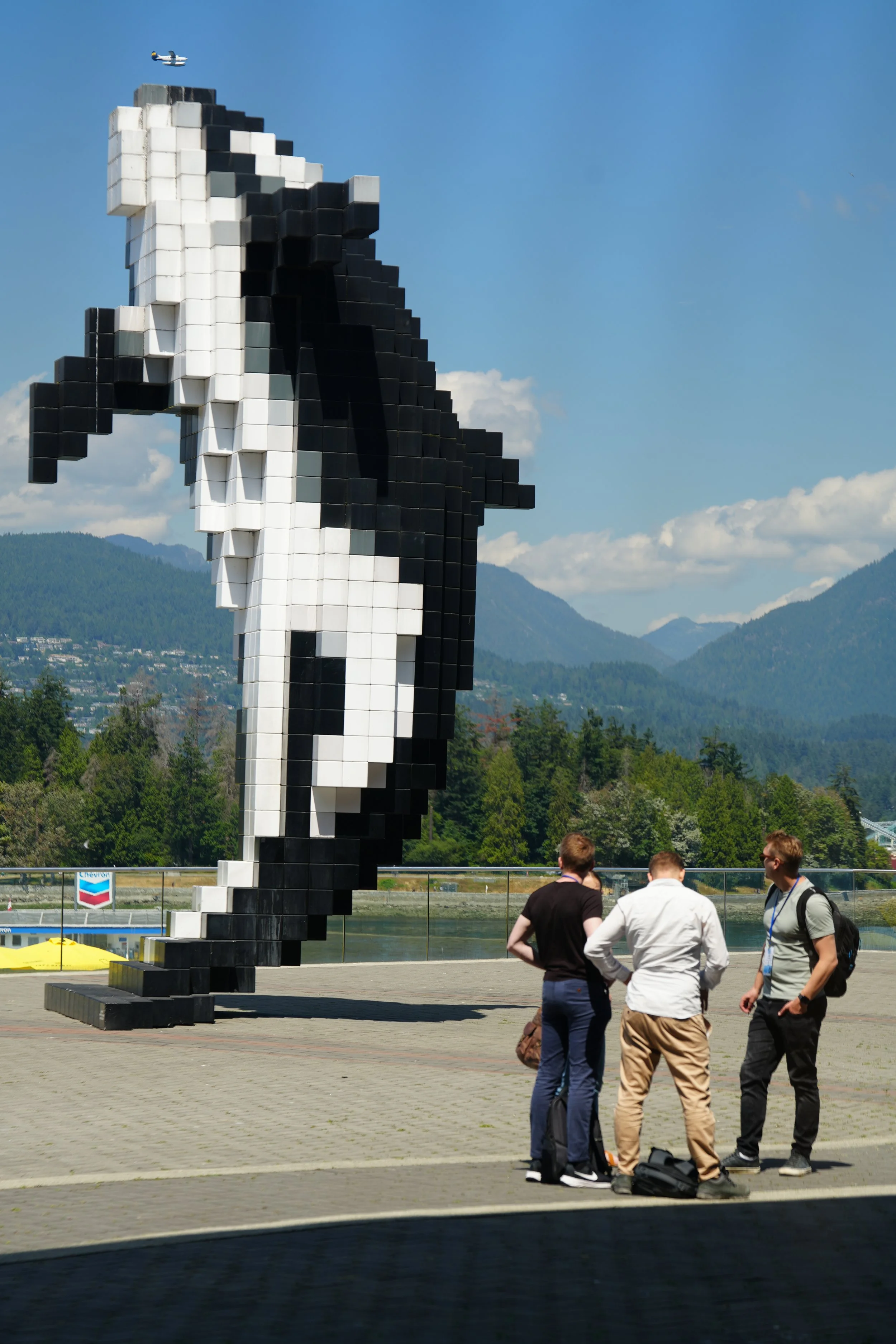 Three people standing and talking in front of a large pixelated sculpture of a white and black figure, with a mountainous landscape and a plane in the sky in the background.