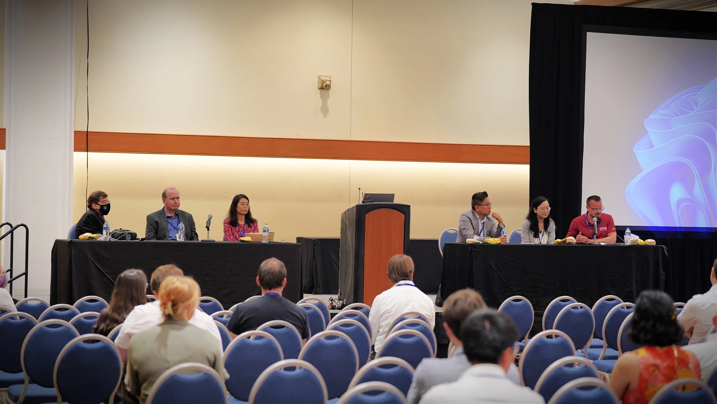 Panel of six people sitting at a long table on a stage, attending a conference or presentation, with an audience in front. The background includes a large projector screen displaying abstract blue shapes.