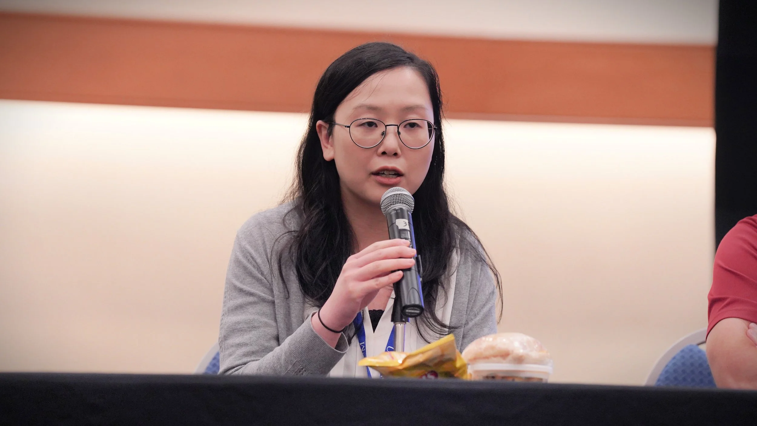 A woman with black hair, glasses, and a gray sweater speaking into a microphone at a panel discussion or conference.