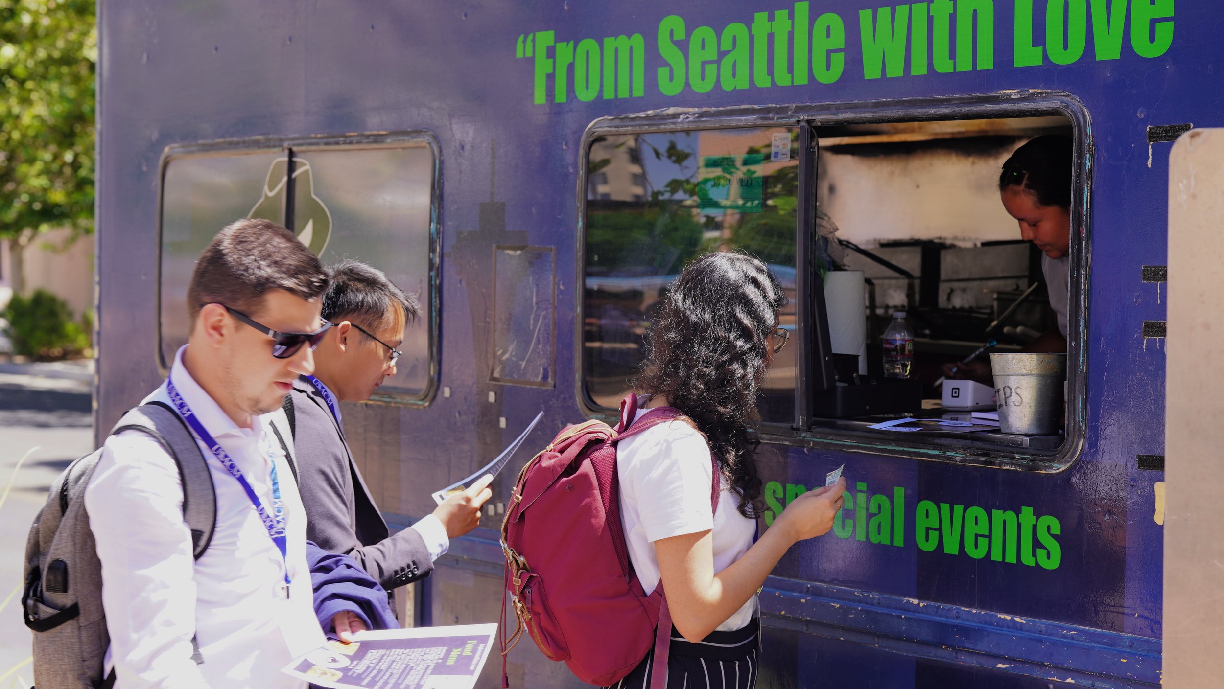 Four people standing in line at a bus ticket window, reading their documents. The bus window is blue with green text that reads, 'From Seattle with Love' and 'special events.' A woman with long curly hair and a maroon backpack is at the window, purchasing a ticket or asking for information.