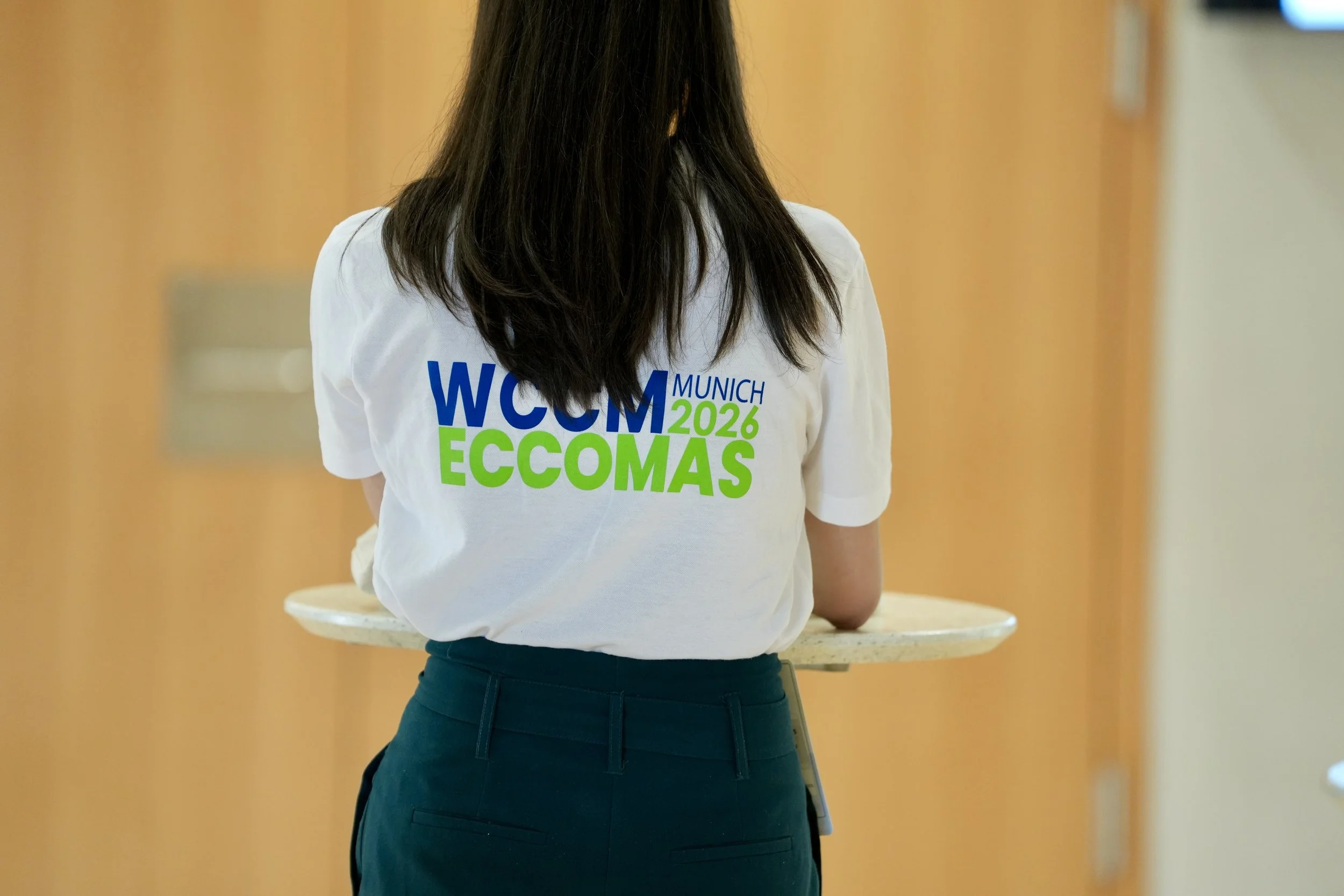 Person with long dark hair wearing a white T-shirt with colorful text on the back, standing in front of a yellow wall, holding a round table.